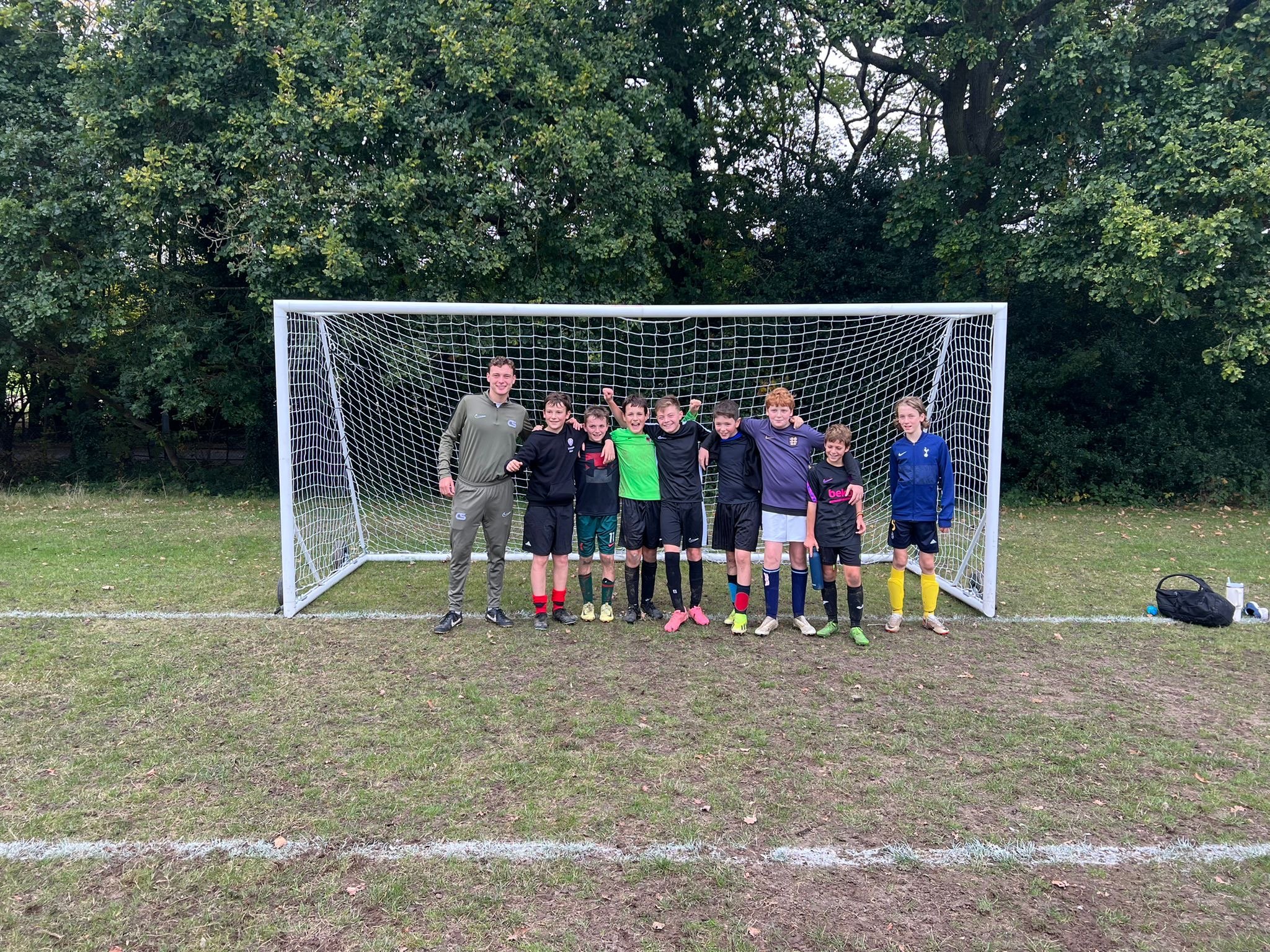 Group of young football players posing with a coach in front of a goal on a grassy field.