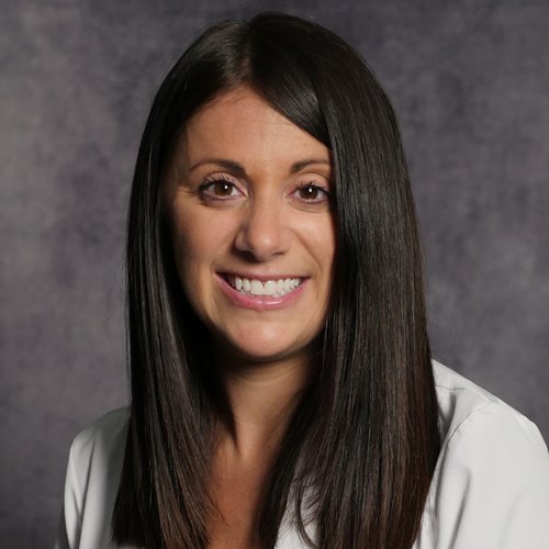 A woman with long straight black hair smiling at the camera, wearing a white blouse, against a dark gray background.
