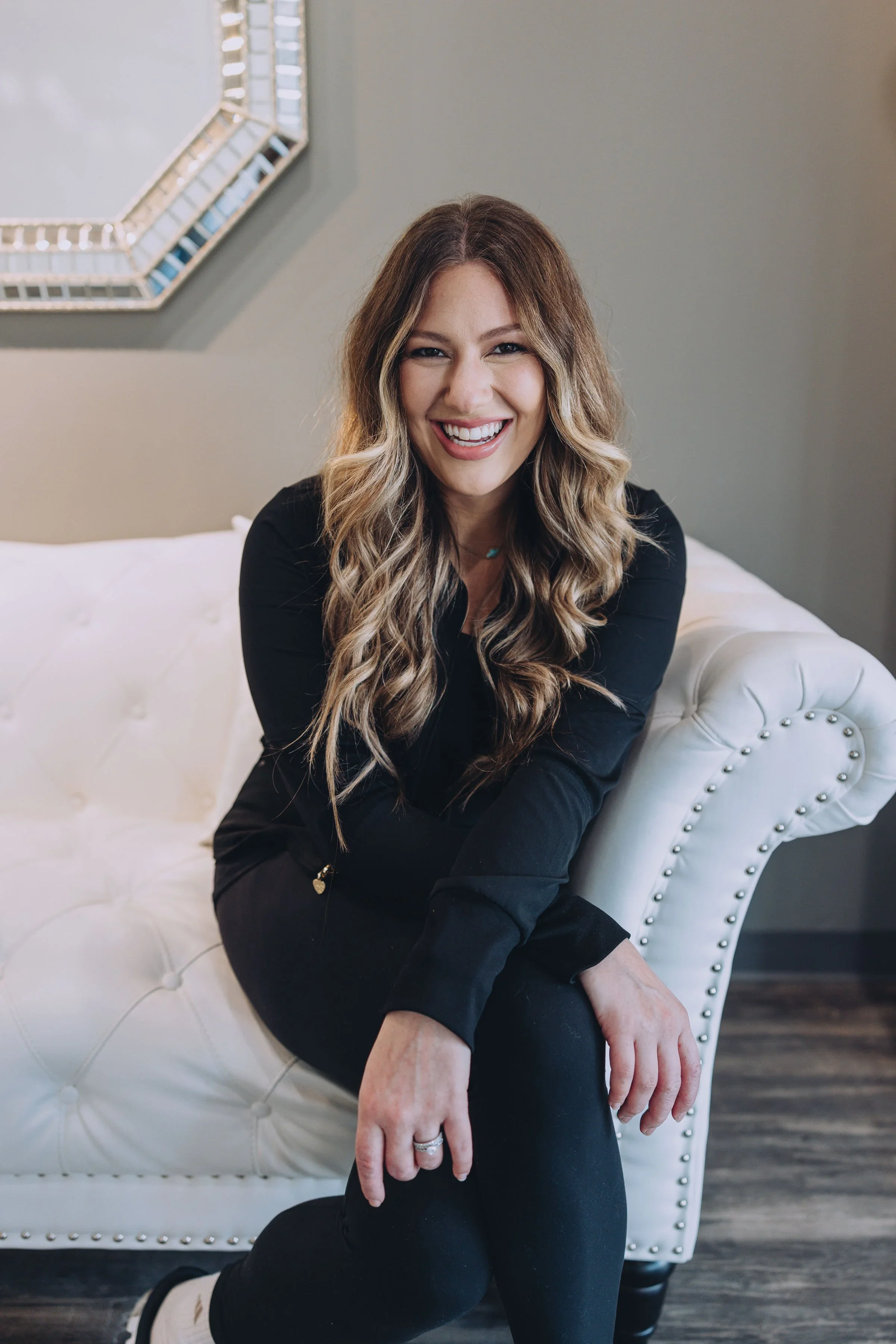 A smiling woman with long wavy blonde hair, wearing a black top and black pants, sitting on a white upholstered chair with nailhead trim, in a room with gray walls and a decorative mirror.