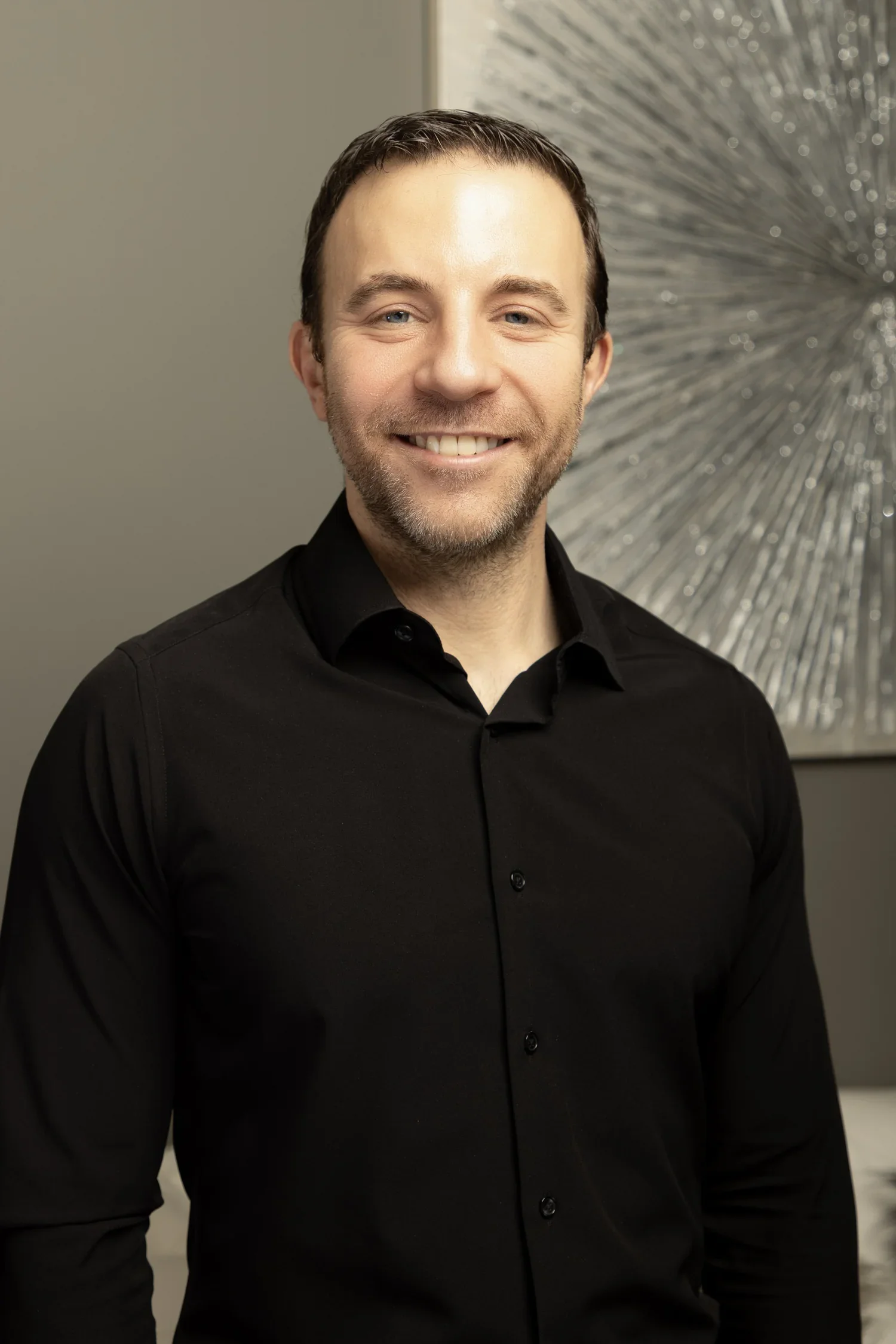 Headshot of a smiling man with short dark hair, a beard, wearing a black shirt, in front of abstract silver wall art.