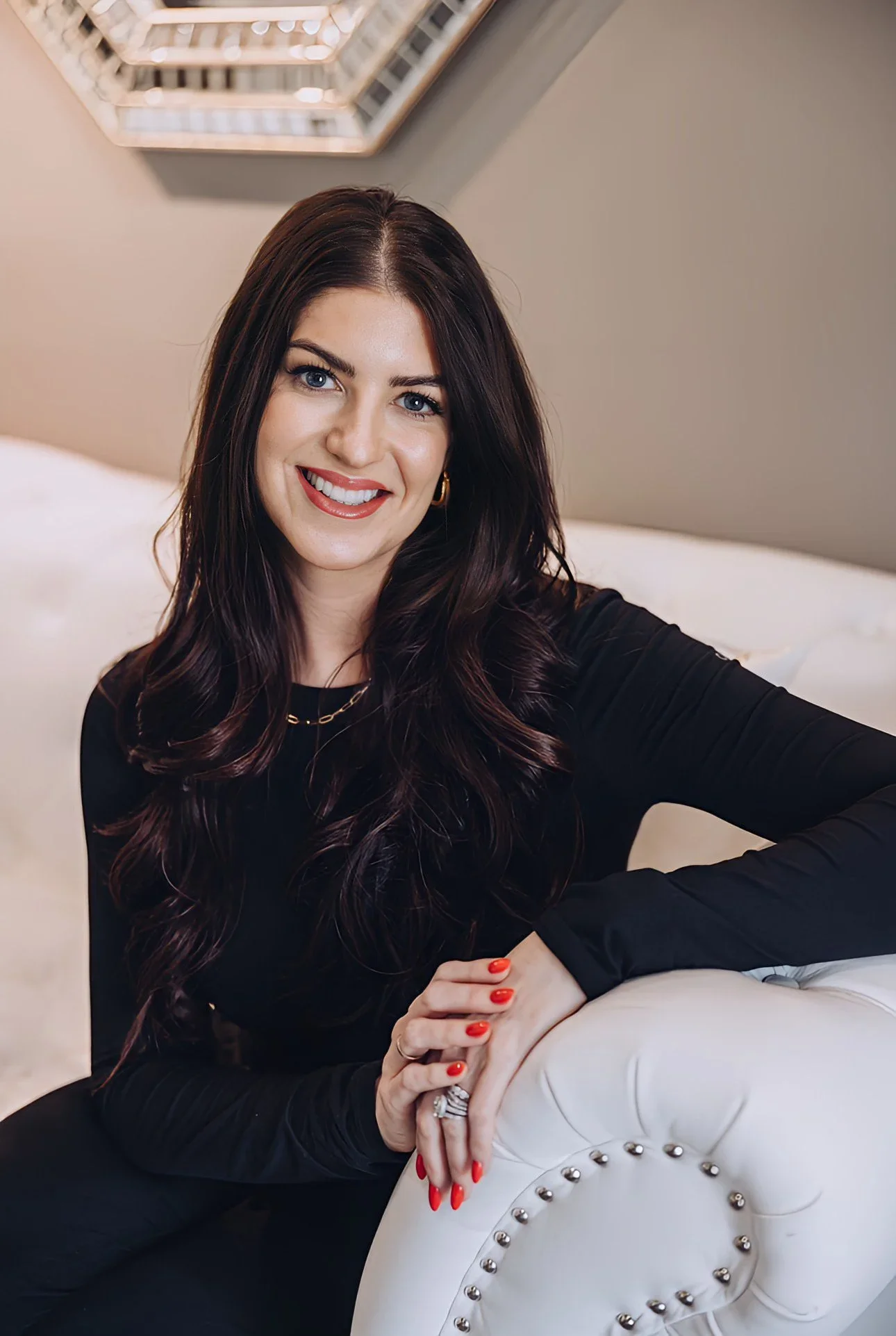 A woman with long dark wavy hair, wearing a black long sleeve top, smiling and taking a selfie inside a modern, well-lit room with a beige couch and a geometric wall decoration.