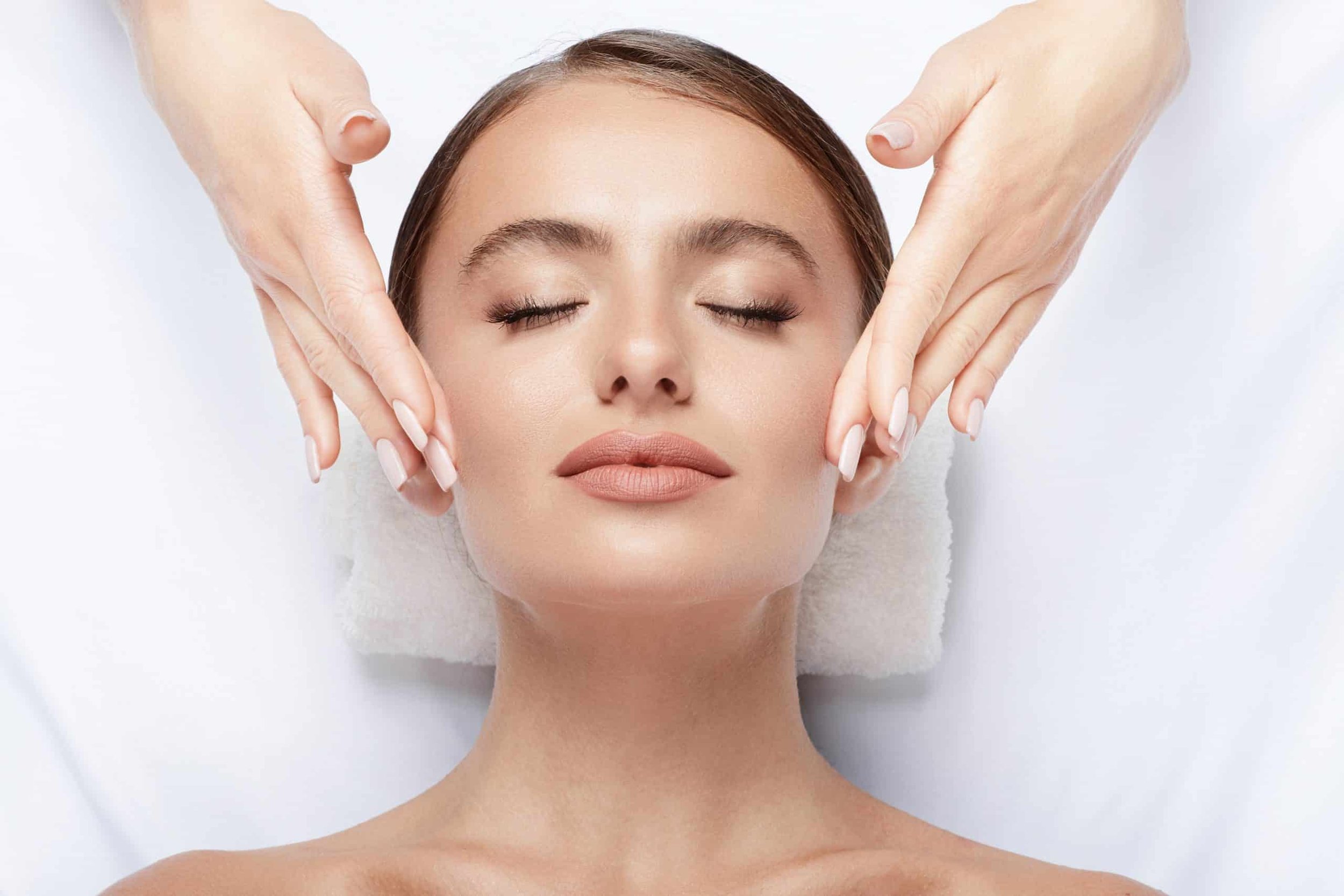 A woman receiving a facial massage or treatment, lying down with eyes closed, with a serene expression.