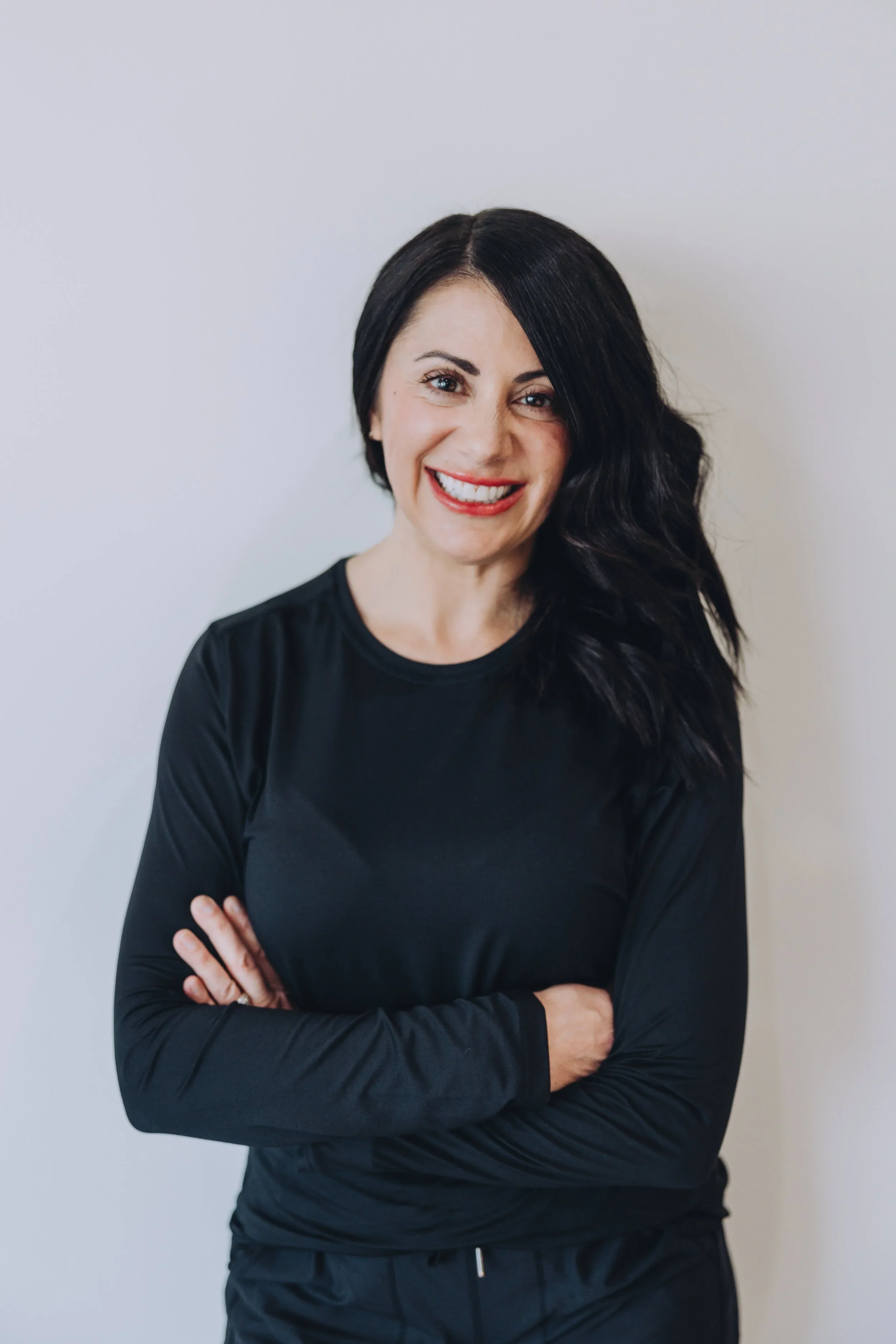 A woman with long, dark, wavy hair smiling with arms crossed, wearing a black long-sleeve shirt, standing against a plain light gray background.