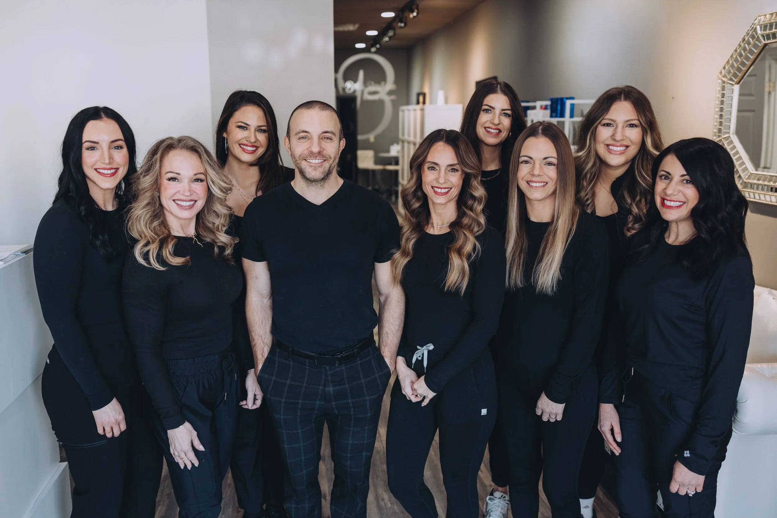 Group of nine smiling people standing together indoors, dressed in black clothing, in a modern setting with a mirror on the wall.