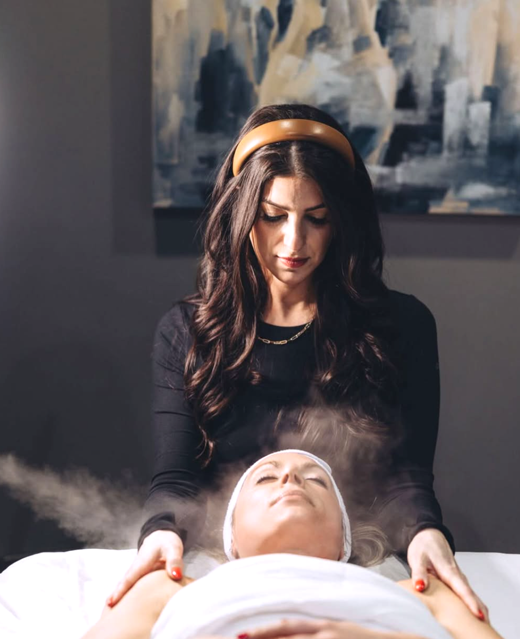 Woman getting a facial treatment at a spa, with steam being applied, while another woman, possibly a therapist, gently holds her shoulders.