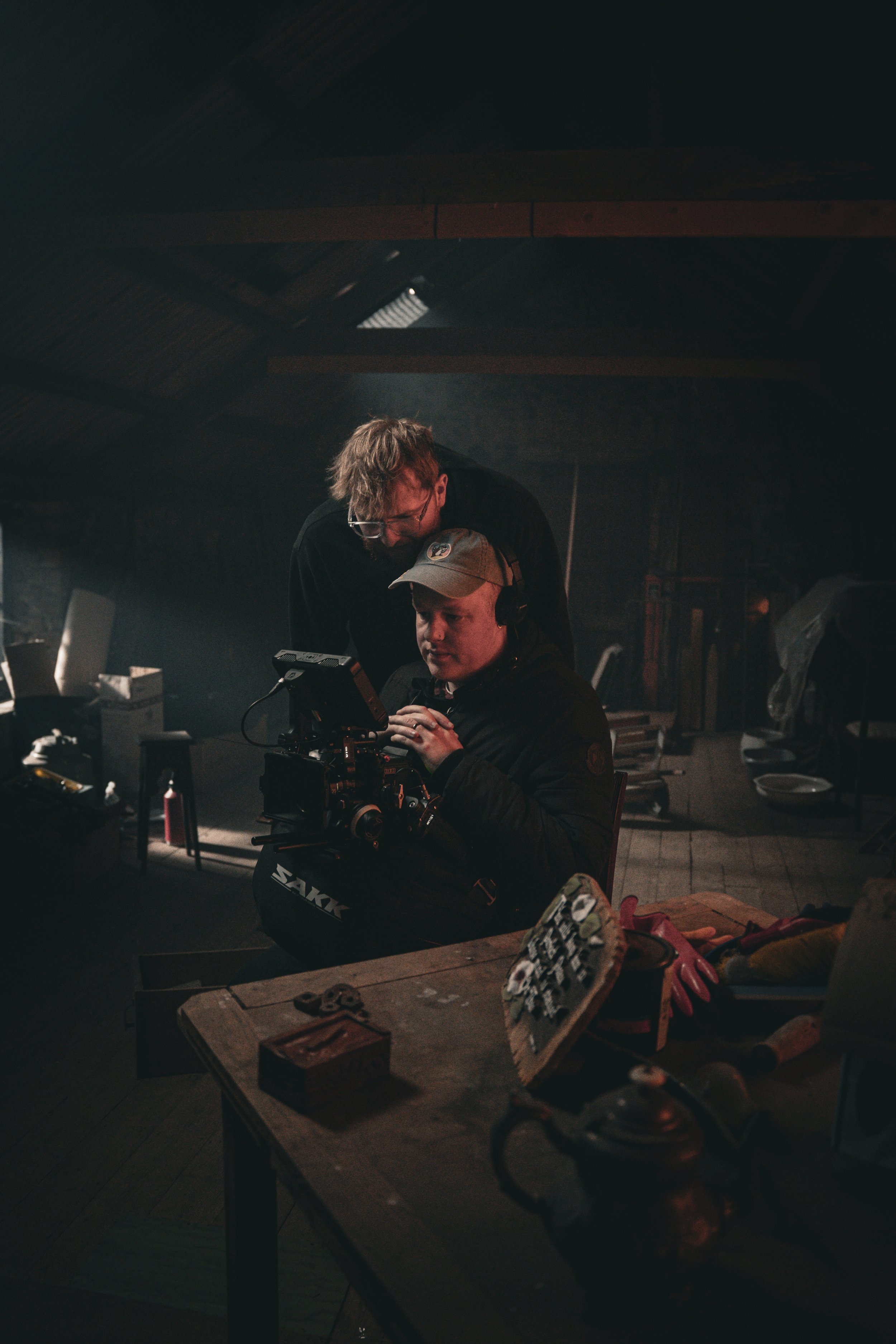 Two men working with professional camera equipment in a dimly lit room, surrounded by various tools and supplies on a cluttered wooden table.