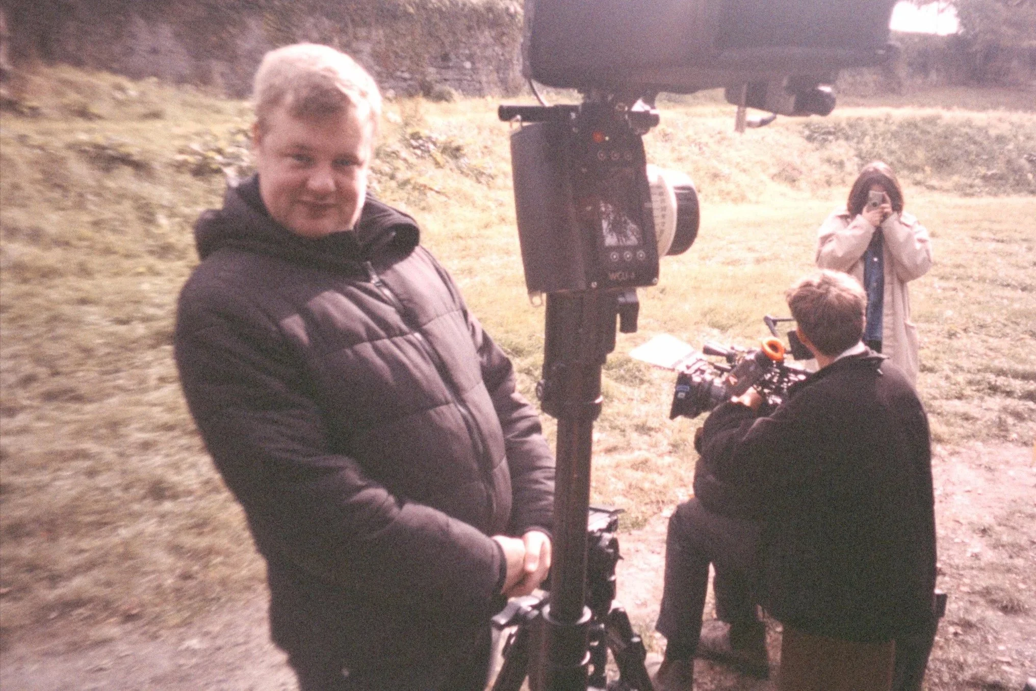 A group of people outdoors near a large camera or binoculars mounted on a tripod, with a young man in a black jacket smiling at the camera and a woman taking a photo with her phone.