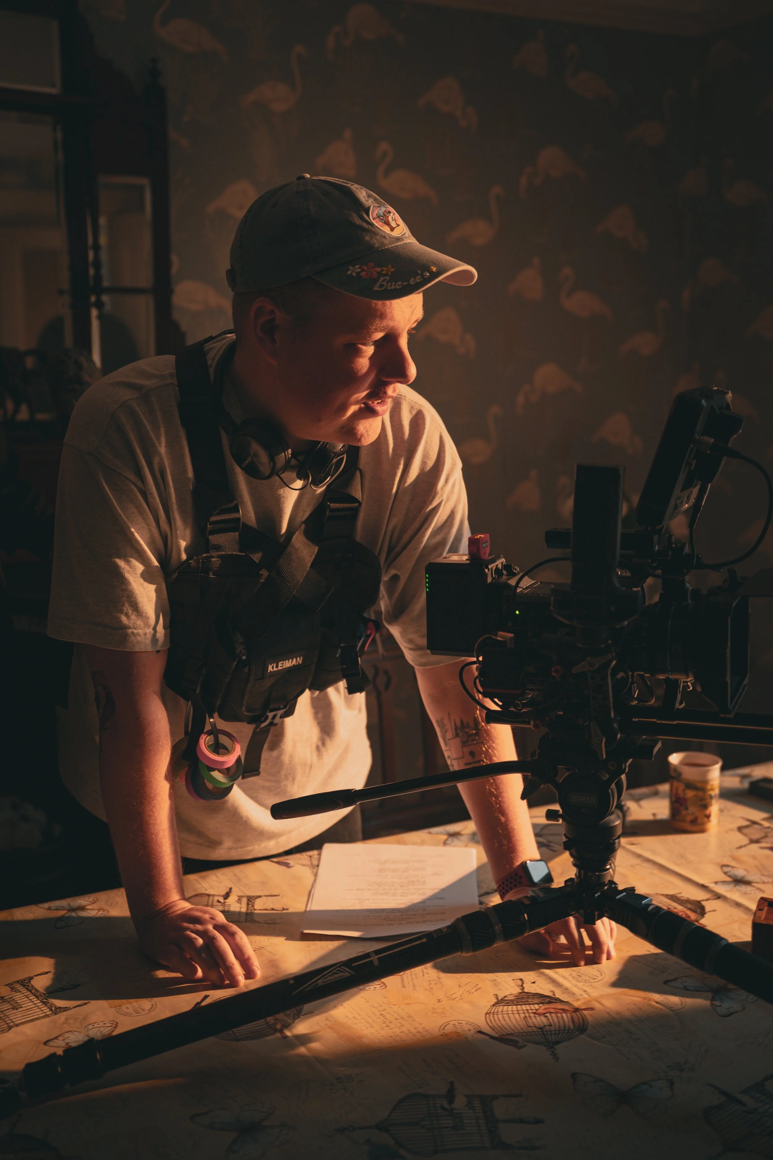 A cinematographer wearing a chest rig and headset looking at a professional cinema camera monitor in a dimly lit room with warm lighting.