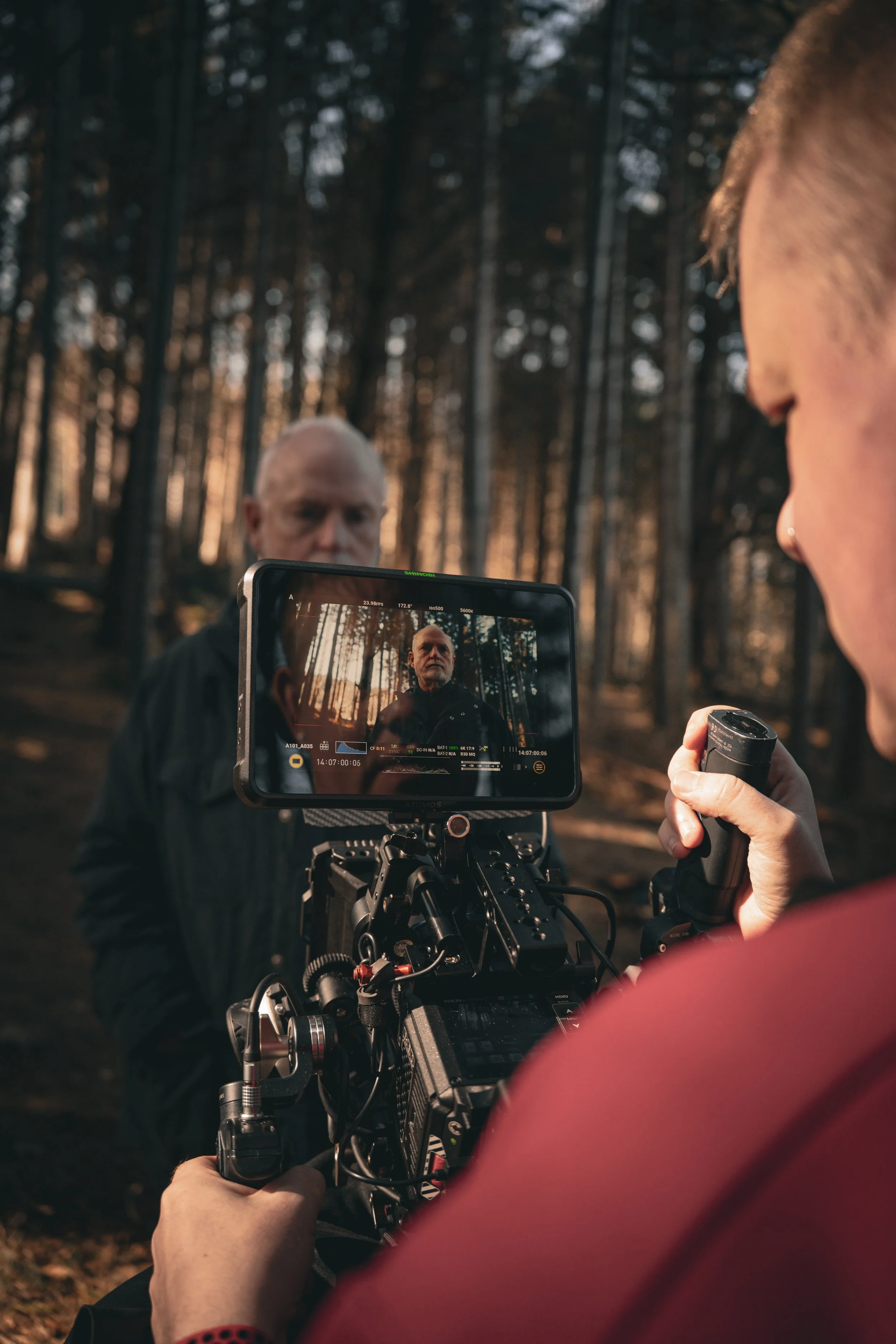 A film crew captures footage of a bald man with a beard in a wooded area during sunset. The photo shows the scene through the camera monitor, with a woman in the foreground holding a camera and a microphone, focused on the man.
