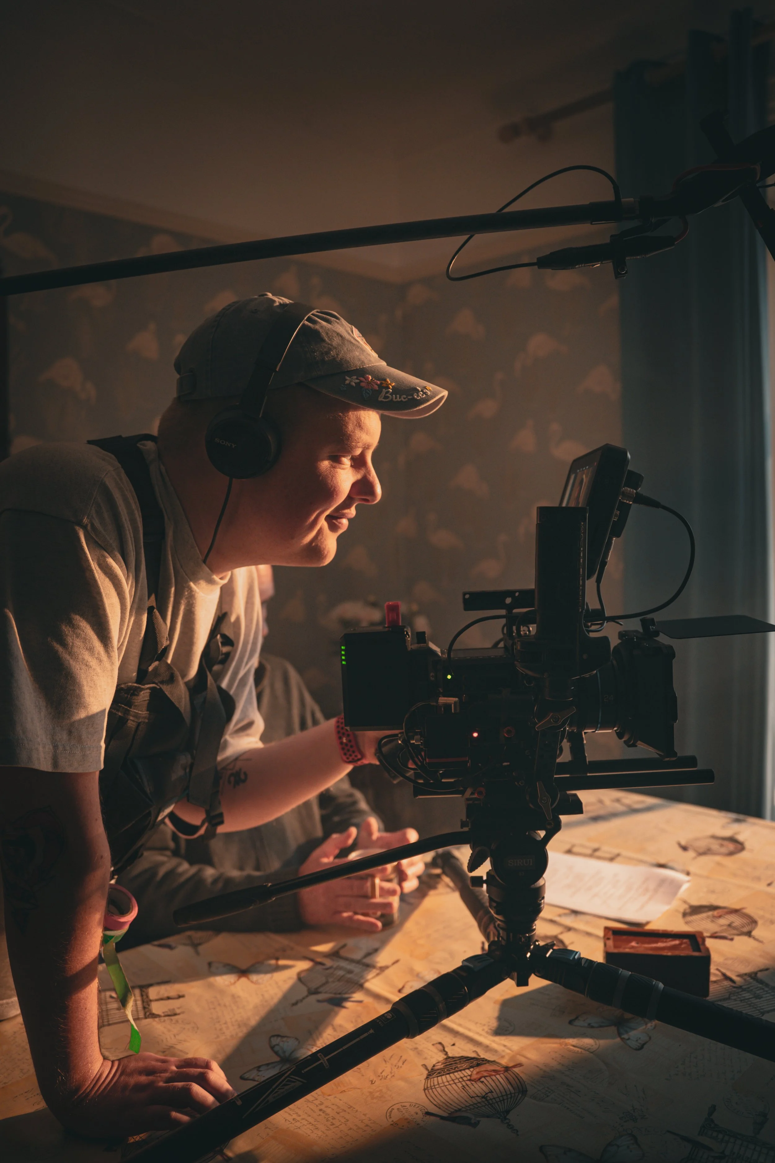 A person wearing a cap with floral embroidery and headphones is looking at a monitor while operating a camera mounted on a tripod. They are in a room with a wooden table covered with vintage-style drawings and maps, illuminated by warm lighting.