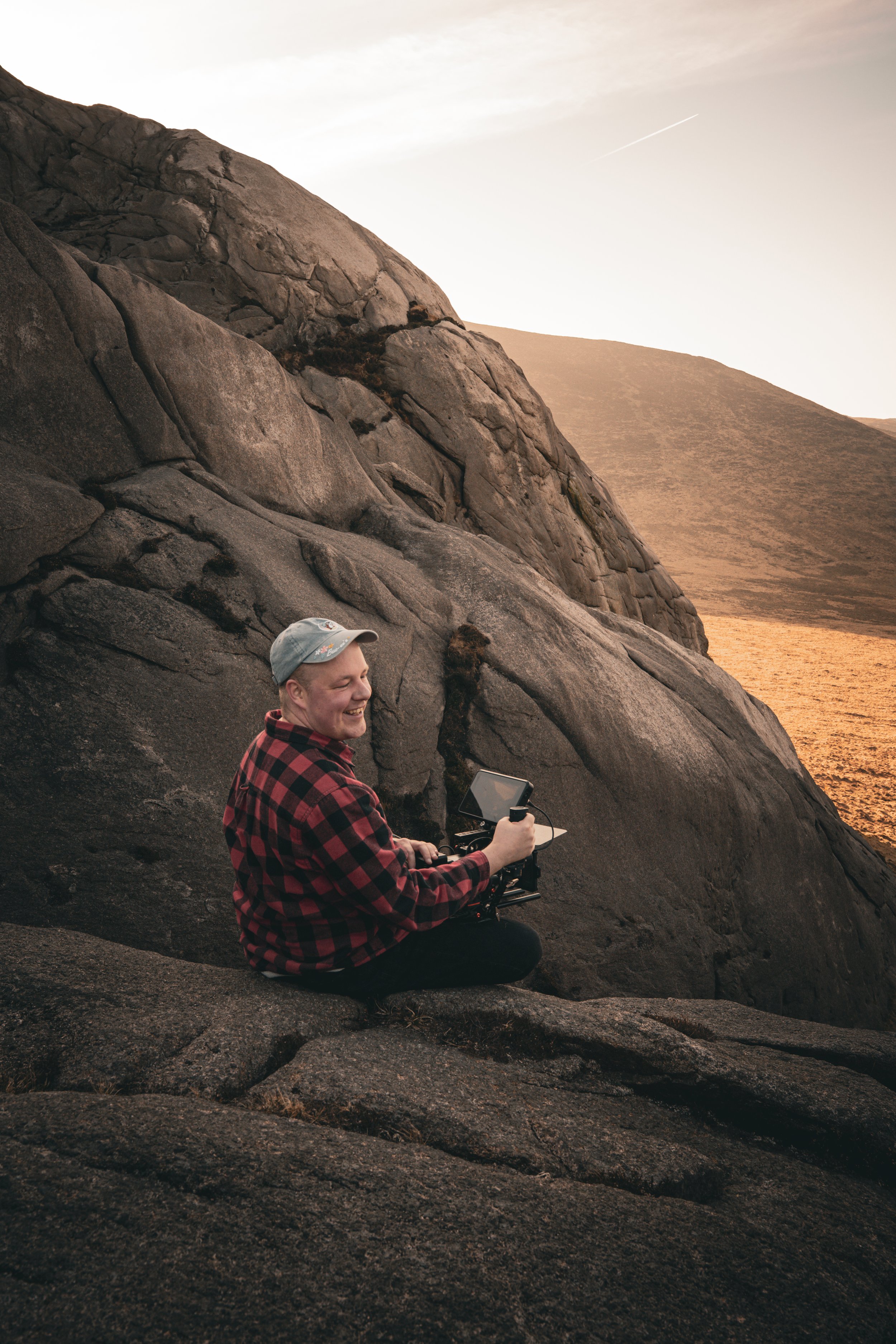 A cheerful man in a red and black plaid shirt and a gray cap is sitting on a rocky landscape in front of large brown rocks, holding what appears to be a camera stabilizer or drone controller, with the sunset or sunrise casting a warm glow on the dry 