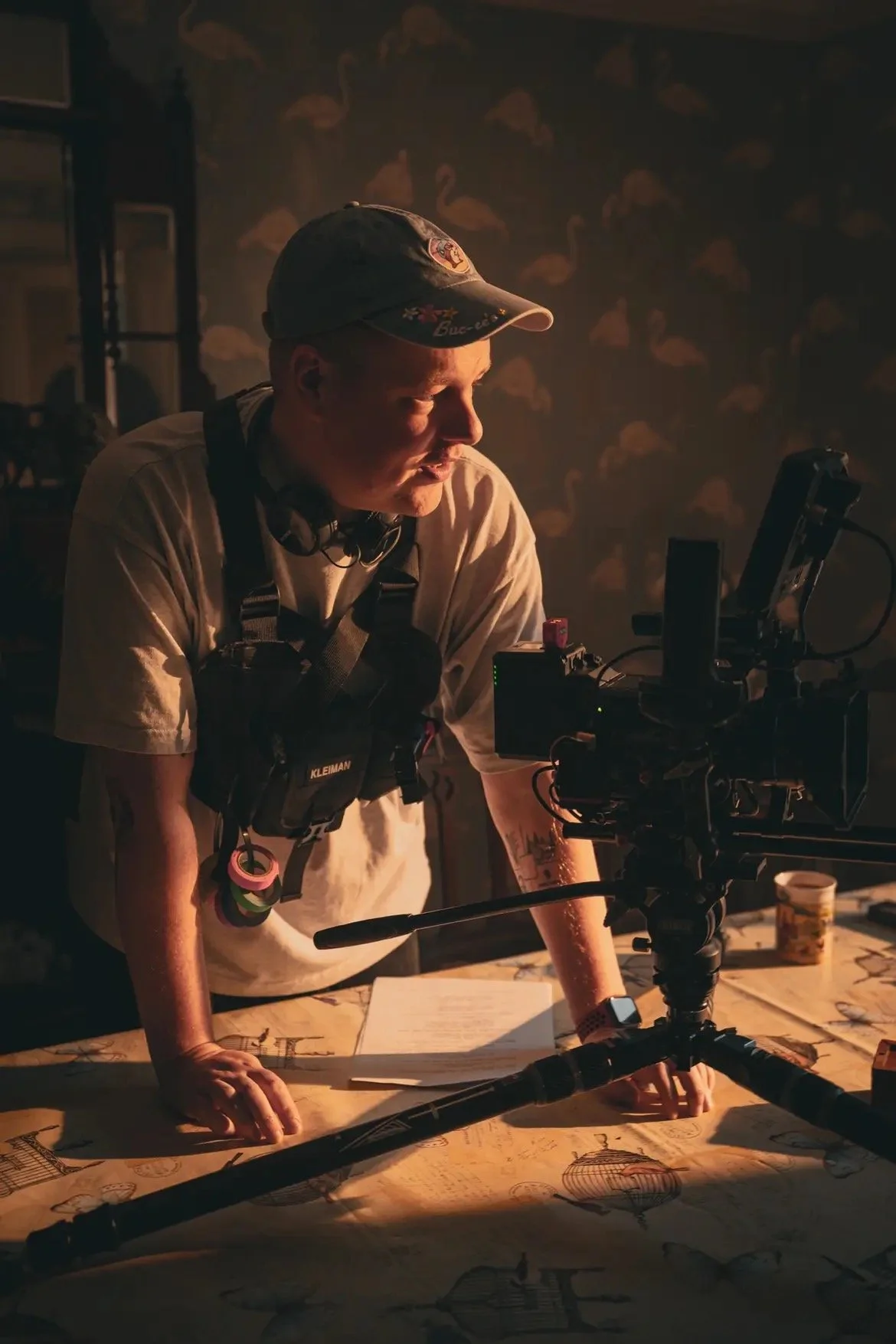 A person operating camera equipment and looking at documents on a table, with a dark background decorated with swan patterns.