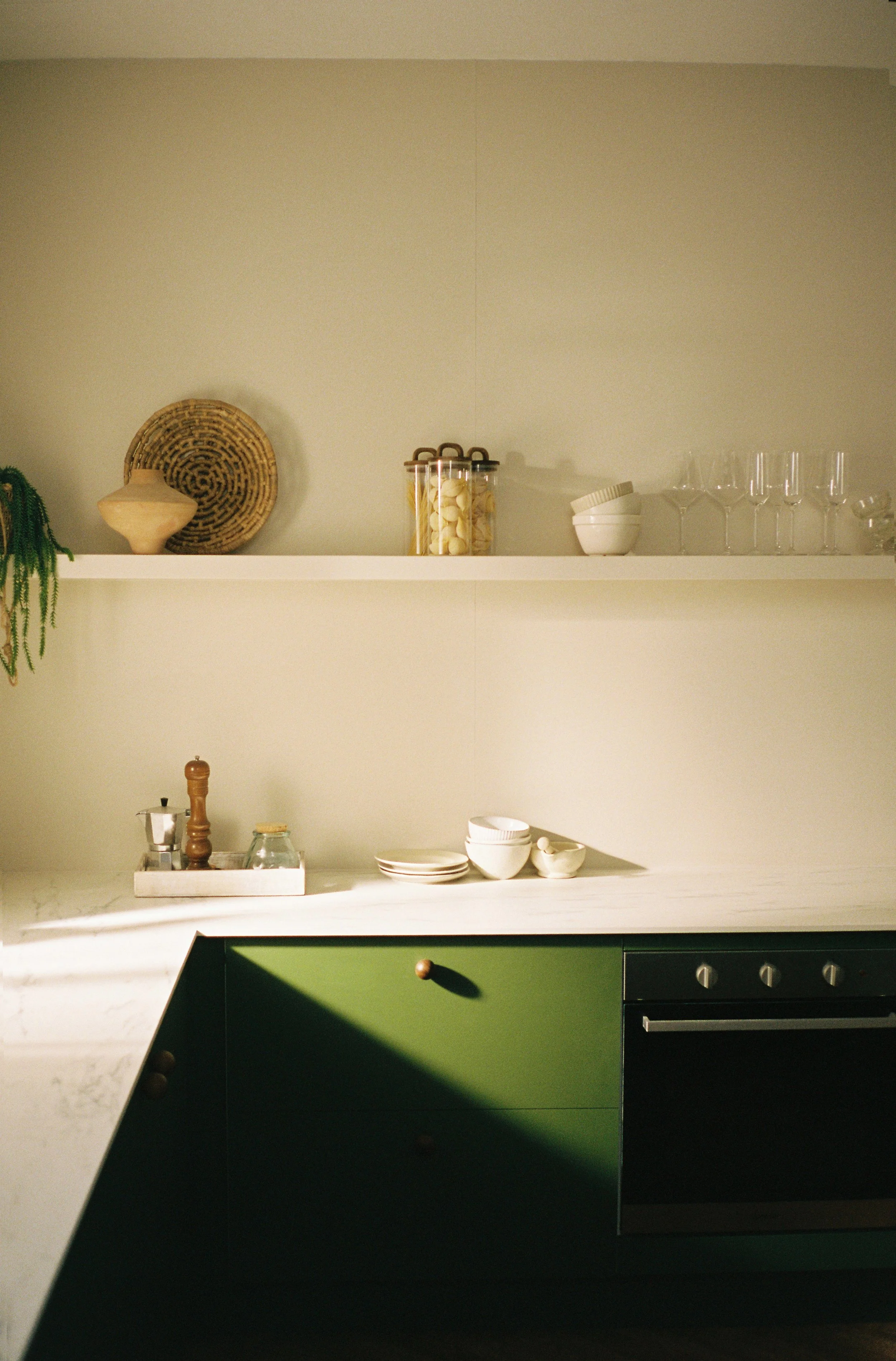 A modern kitchen with a white countertop, green cabinets, and open shelves displaying decorative bowls, glass jars, and glasses, with sunlight casting shadows. Whisk Studios, kitchen space and film studio, available to hire in Bristol.