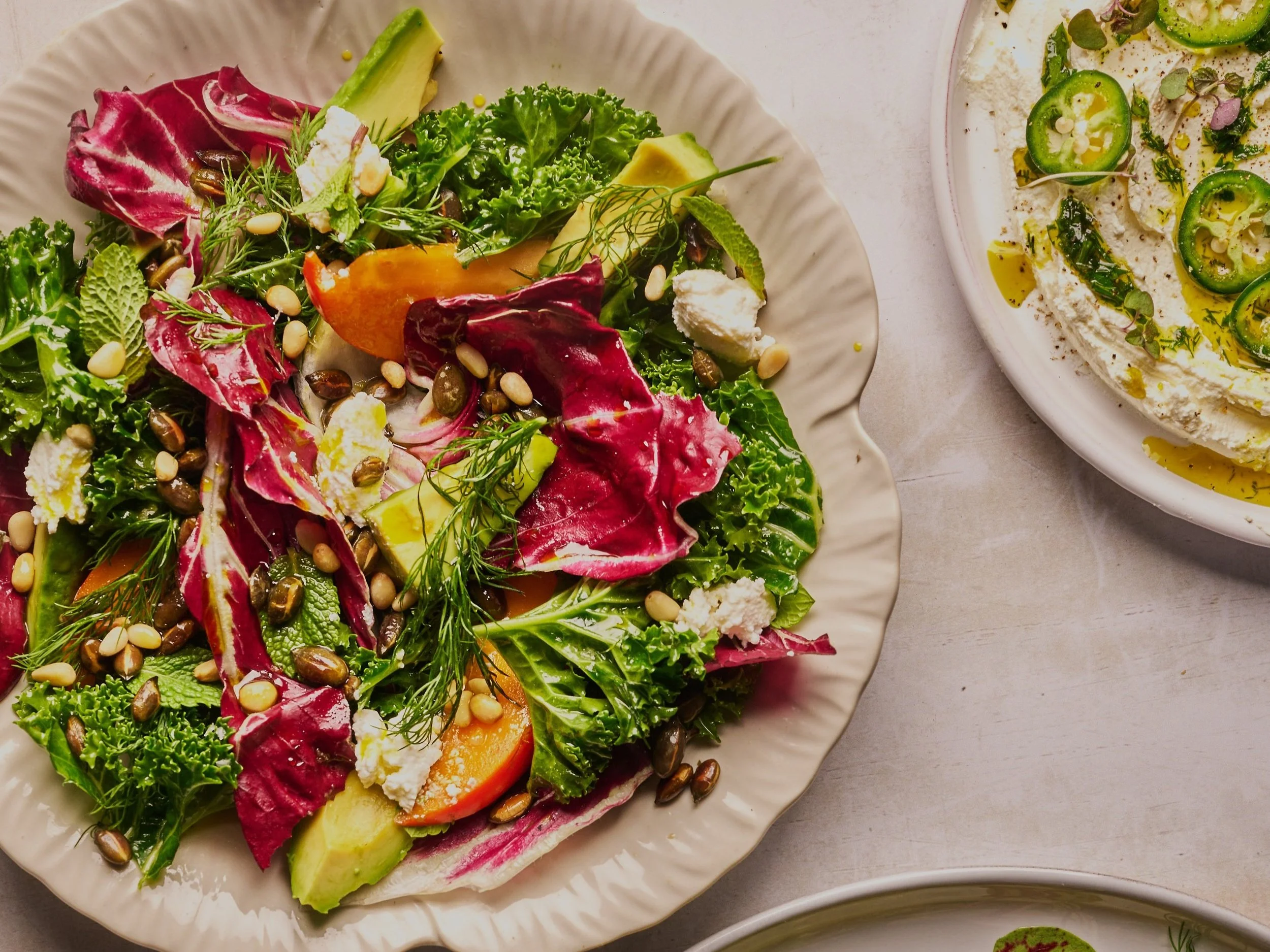 Mixed green salad with lettuce, radicchio, cucumber, carrot, avocado, feta cheese, pine nuts, and fresh herbs on a white plate.