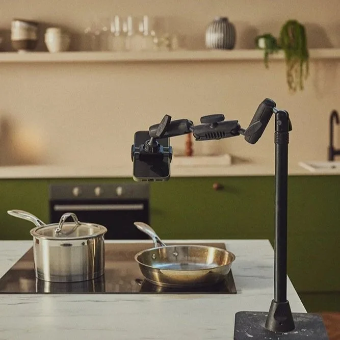 Phone camera tripod over stainless steel frying pan and saucepan on black induction hob. White marble worktop in bright green painted kitchen with inset oven. Neutral painted walls and kitchen shelf with plants, wine glasses, stacks of bowls.