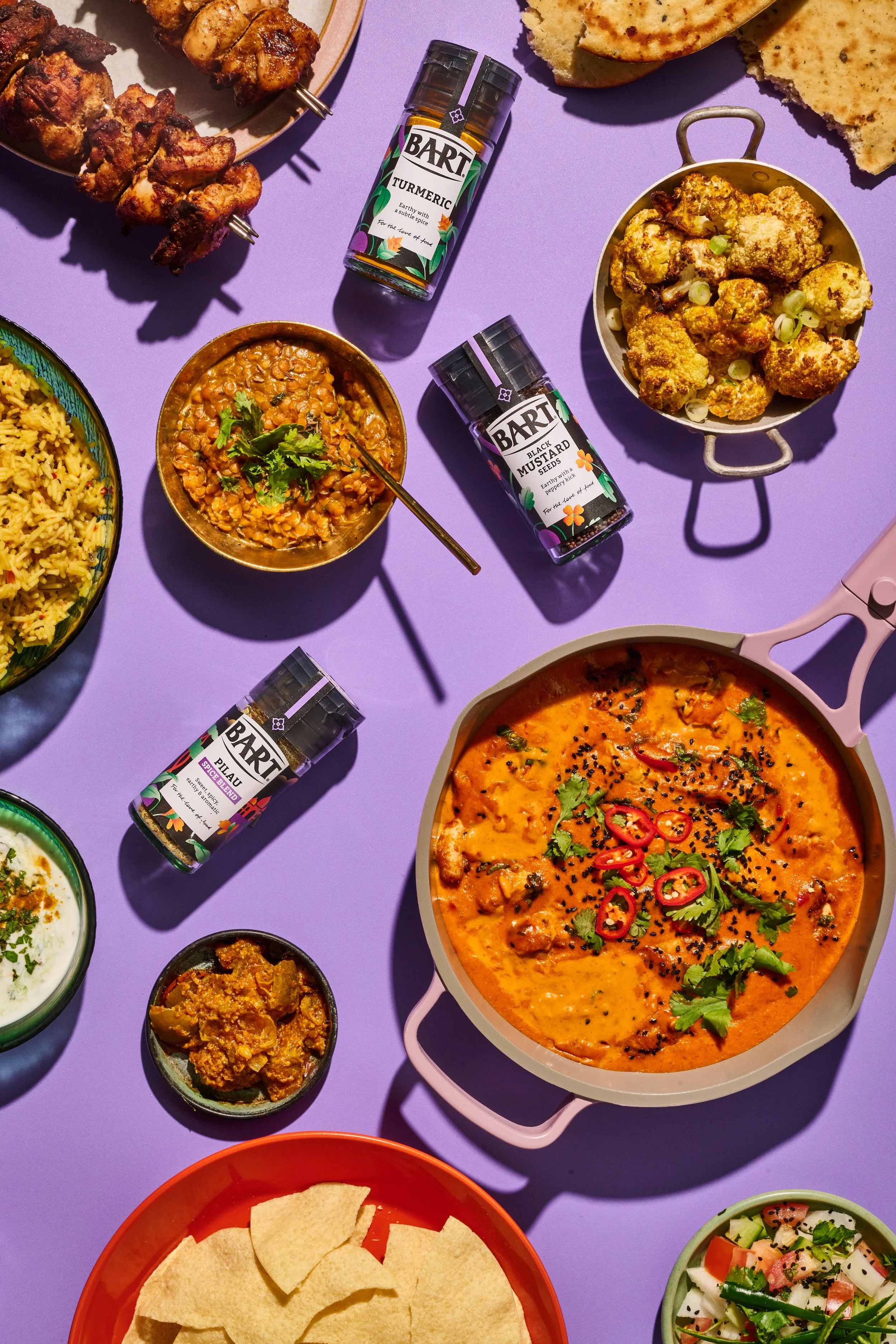 An assortment of Indian dishes including a curry with cilantro, naan bread, fried cauliflower, lentil soup, rice, and other side dishes, along with spice bottles on a purple background.