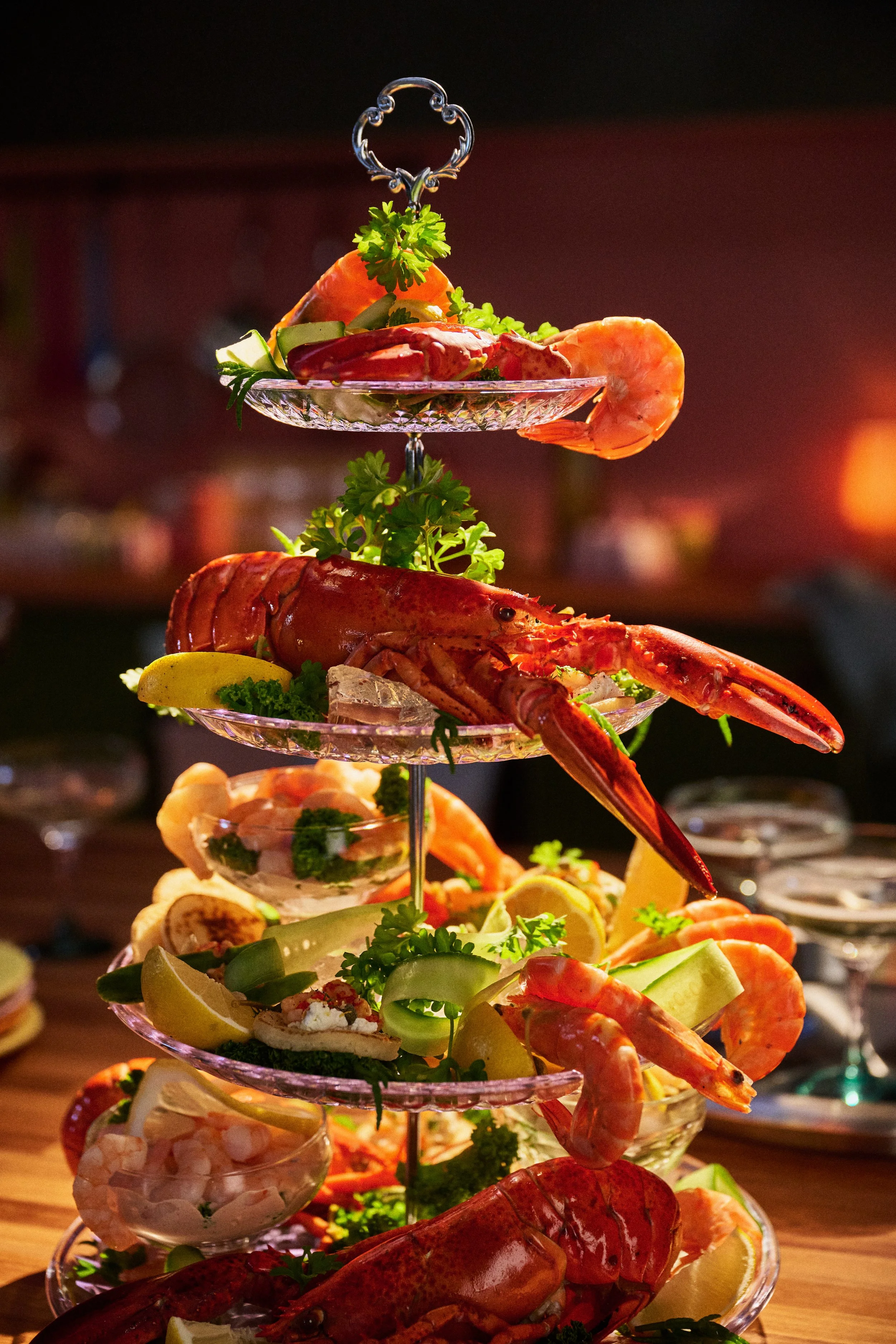 tiered stand of seafood - whole lobster, fresh lettuce, curly parsley, king prawns, tomatoes, blinis, cucumber ribbons in dramatic warm lighting