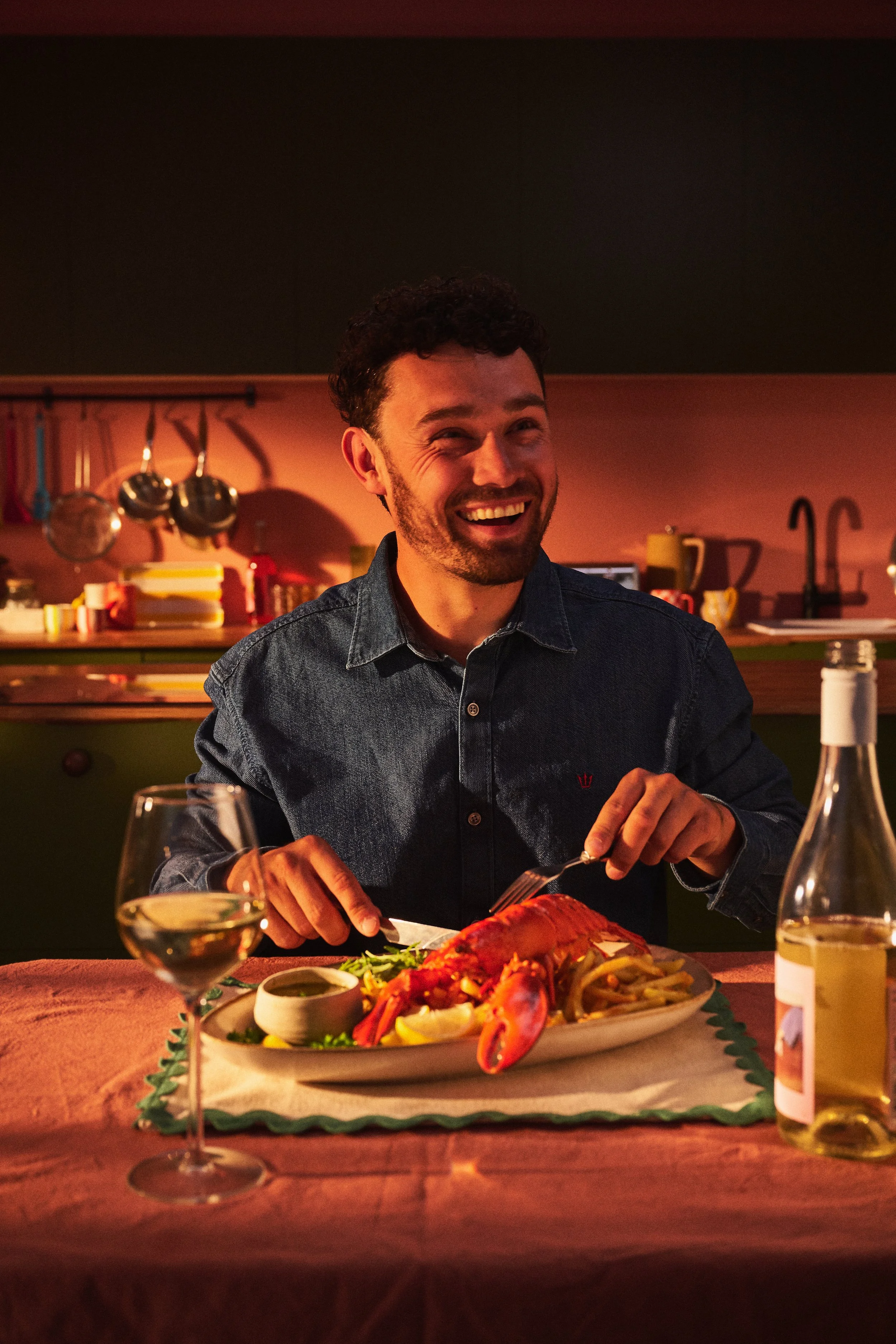smiling man with beard in blue denim shirt, sitting at table with placemat and tablecloth in green and peach kitchen. Large plate of cooked food whole canadian lobster, chips, salad. Bottle and glass of white wine.
