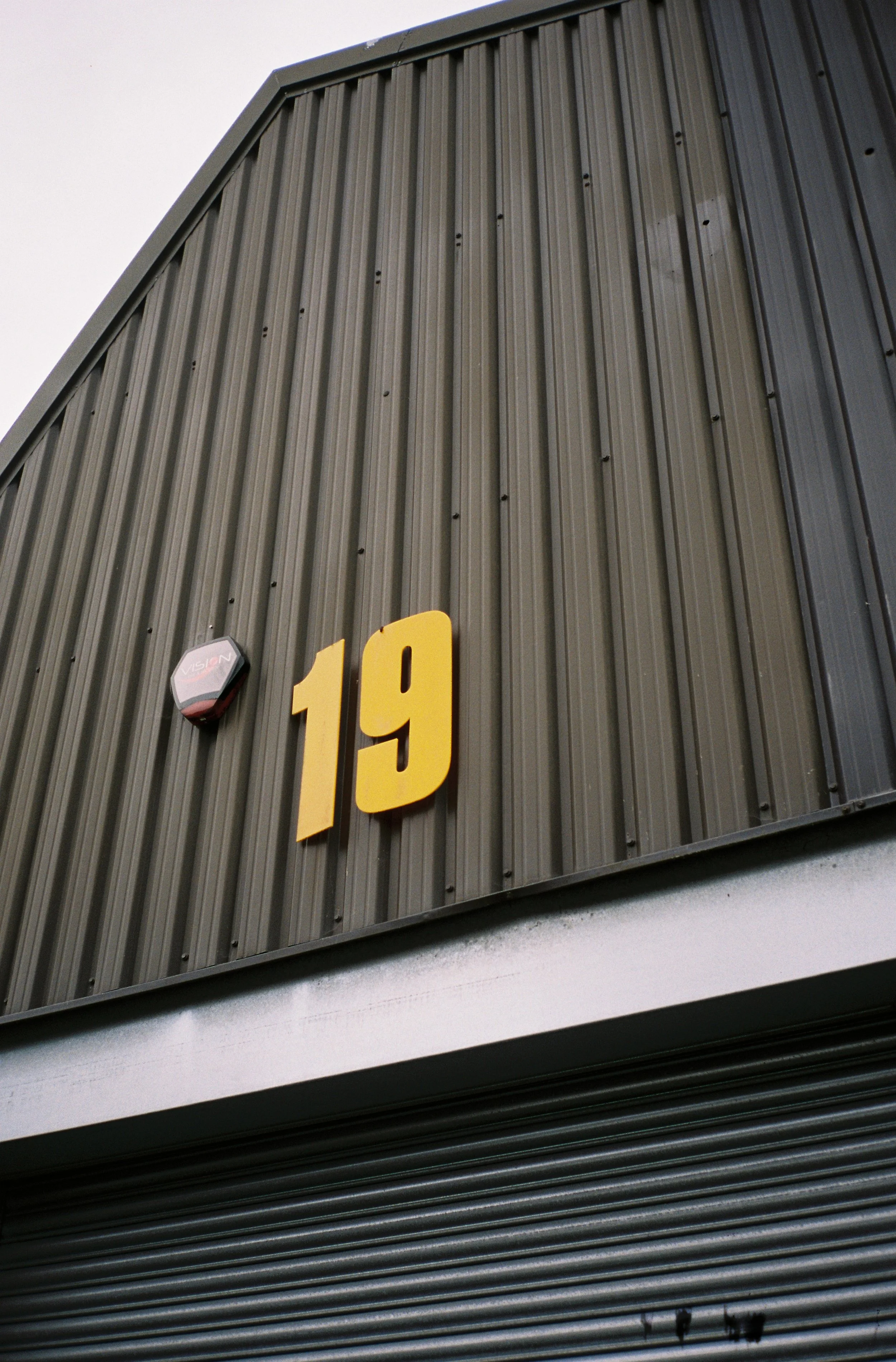 Close-up of a building's exterior with yellow numbers '19' on a dark green corrugated metal wall, a security alarm, and a partially visible roll-up door at the bottom.