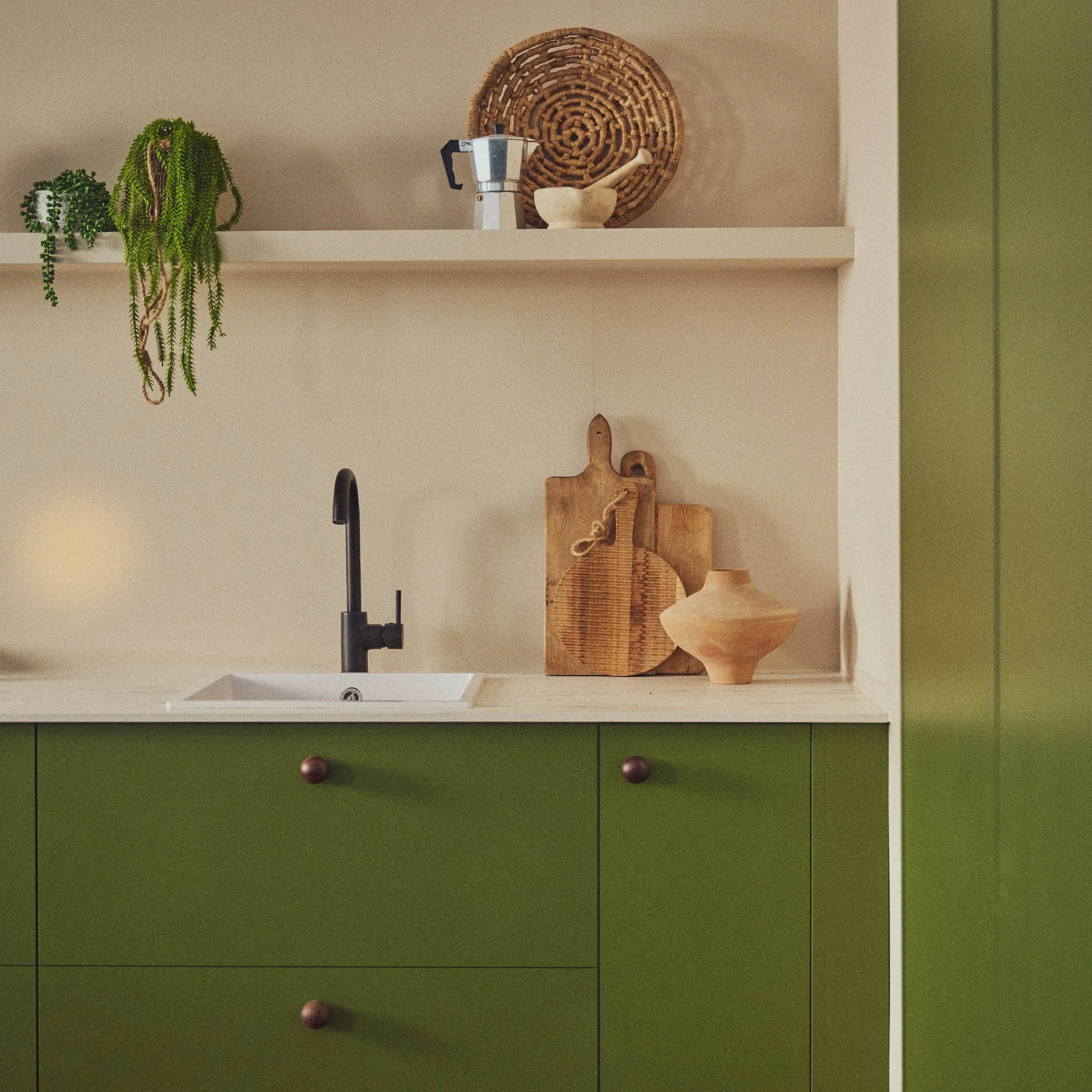 Pea green painted kitchen cabinets with round wooden knobs. Small white sink with black swan neck tap and light marble worktops and shelf with wooden chopping boards, coffee press, plans and pestle and mortar