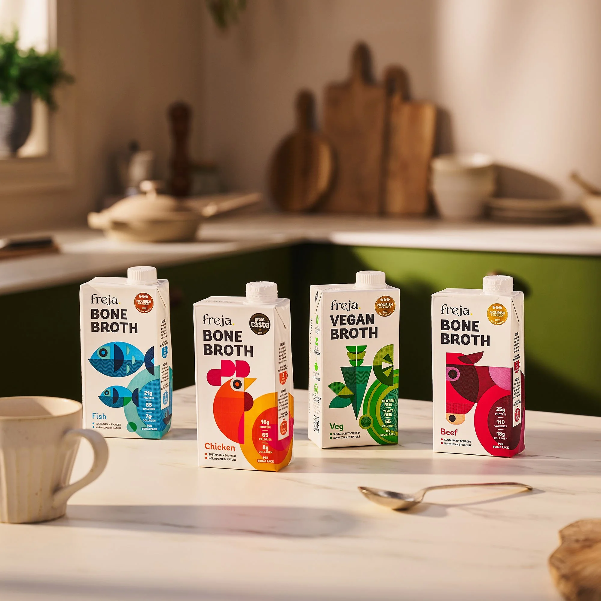 Four Freja Bone Broth cartons in soft lighting on marble kitchen worktop with white cup and stainless steel spoon. Background kitchen worktop with wooden chopping boards, pan and crockery