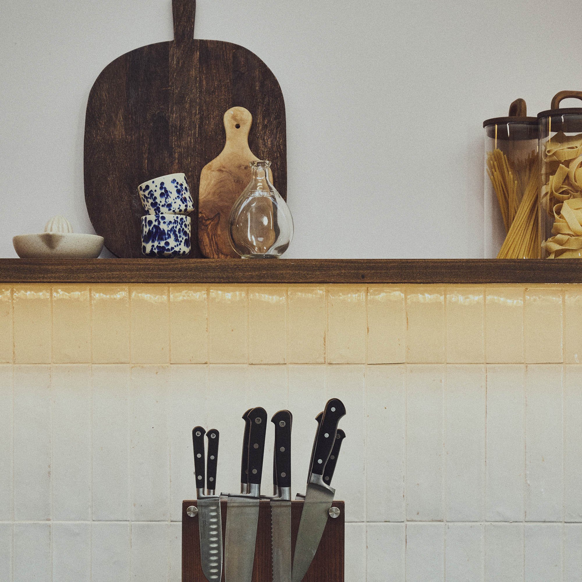 White tiled kitchen wall with knife block of stainless steel knives. LED strip lighting under brown wooden shelf with chopping boards and storage jars 