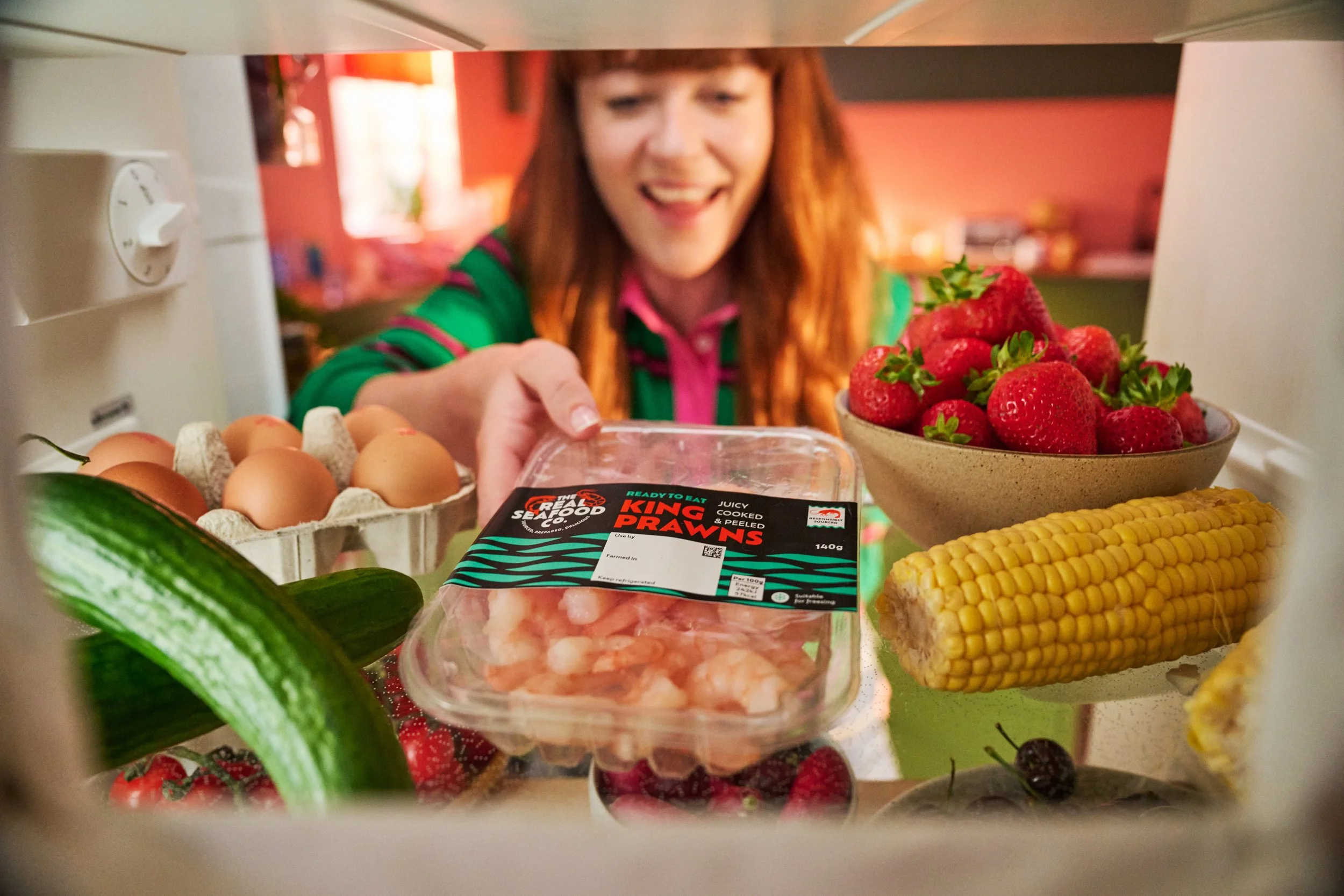 view from inside fridge with open door. Woman reaching for pack of real seafood co king prawns on shelf of fresh, colourful, healthy with fresh cucumber, eggs, sweetcorn on the cob, strawberries, vine tomatoes, cherries.