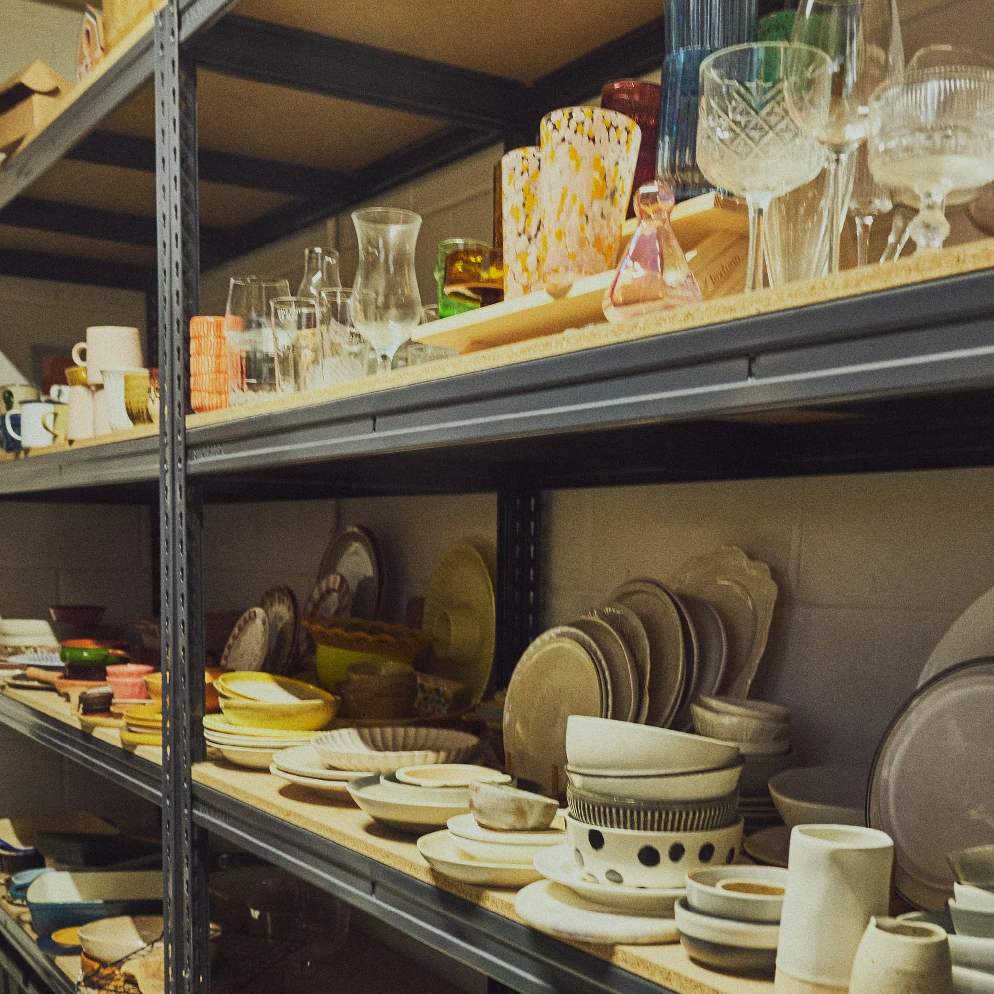 Prop store with metal racking shelves with stacks of white and colourful bowls, plates, wine and drinking glasses 