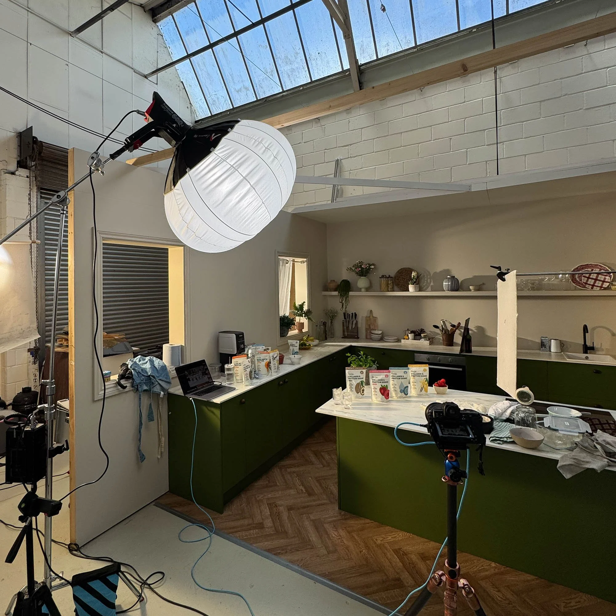 Set kitchen in large industrial spcae with roof lights. Painted pea green green cabinets and island, with light marble worktops and light shelves with serveware. Action shot of photoshoot with camera and lighting kit 