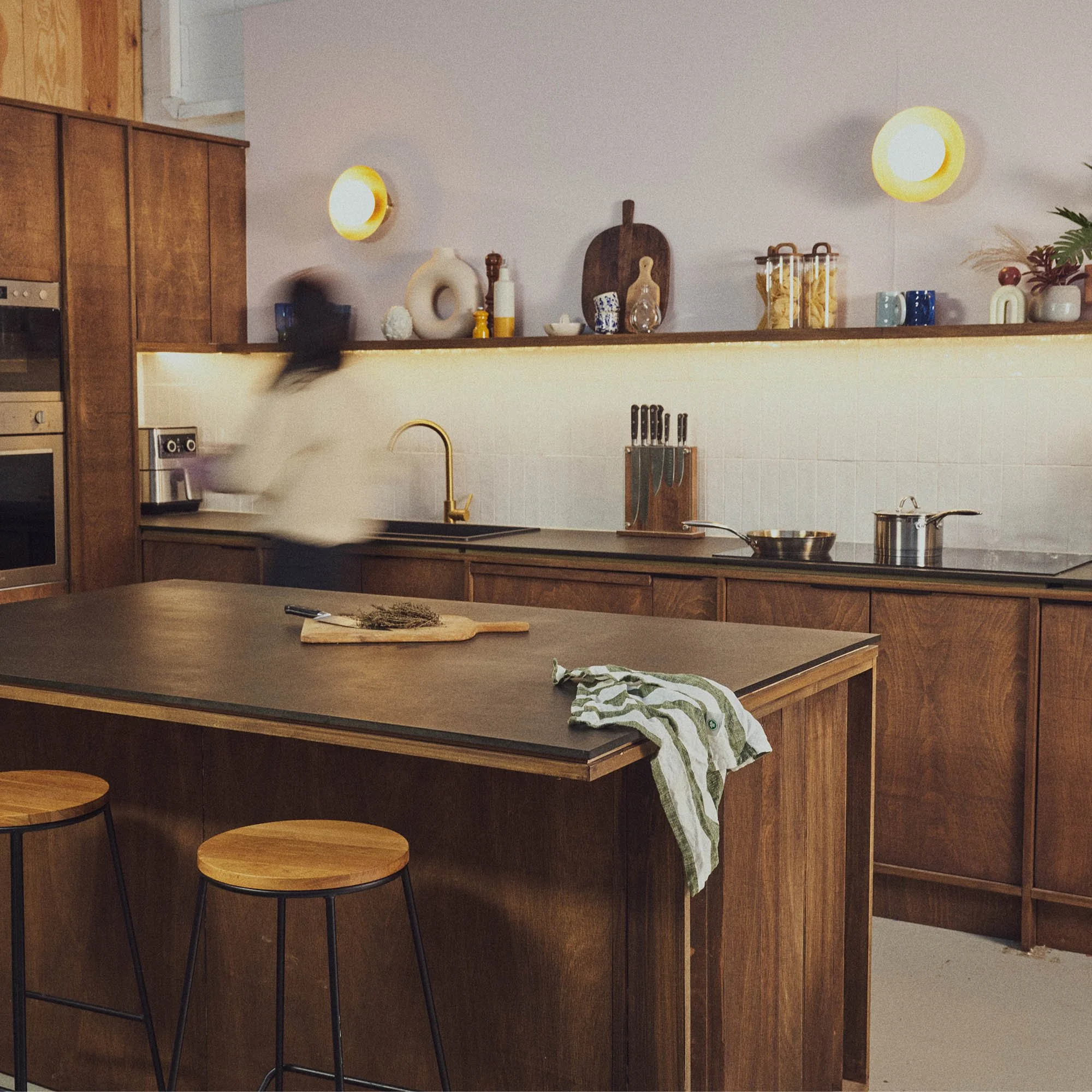 Person moving in dark brown wood kitchen with dark worktops and kitchen island with bar stools. Round wall lights and shelf styled with chopping board, storage jars, vases.