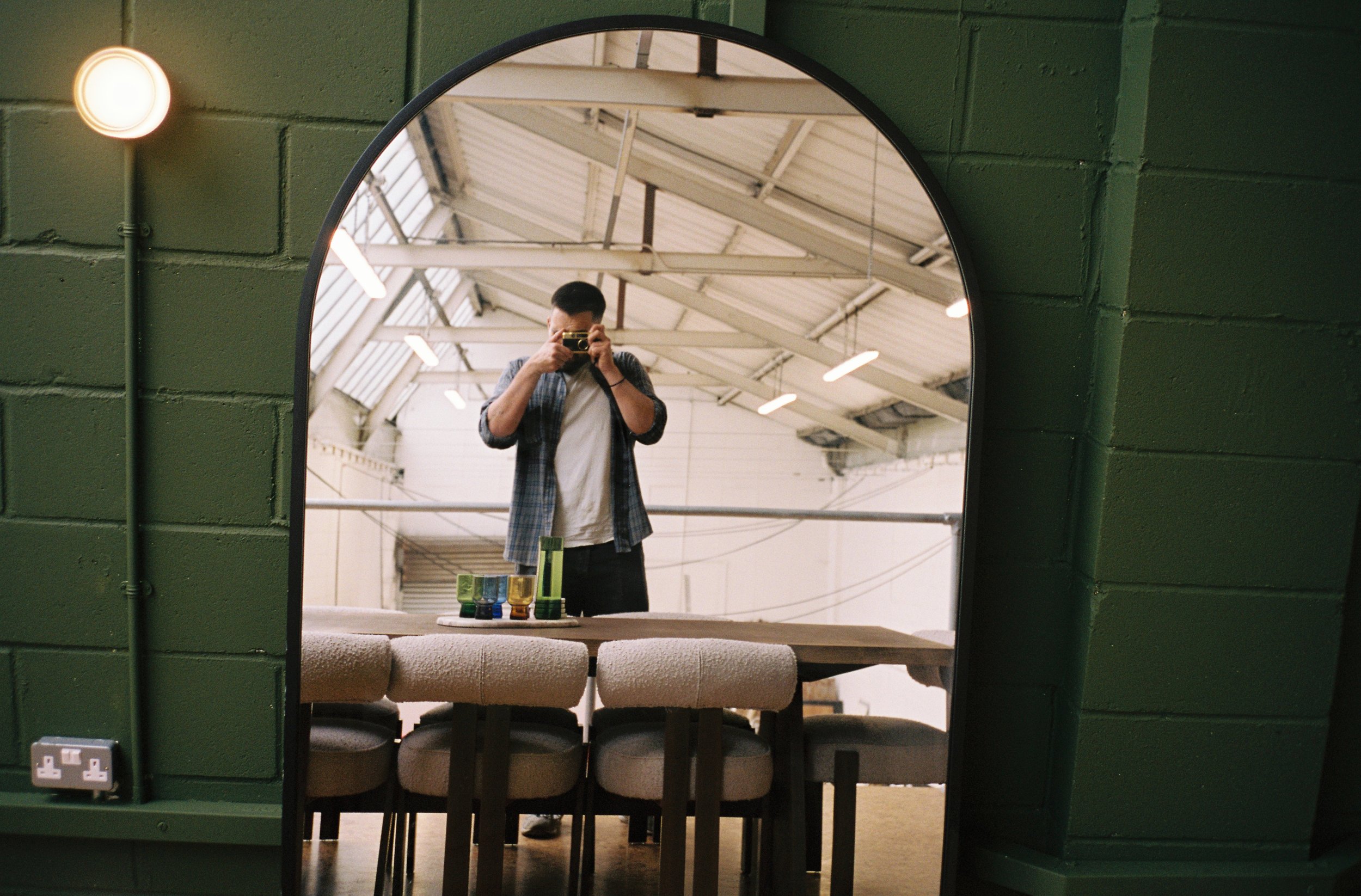 A person taking a photo of their reflection in a mirror, with a large table and chairs visible in the background. The space has an industrial look with high ceilings and exposed beams.