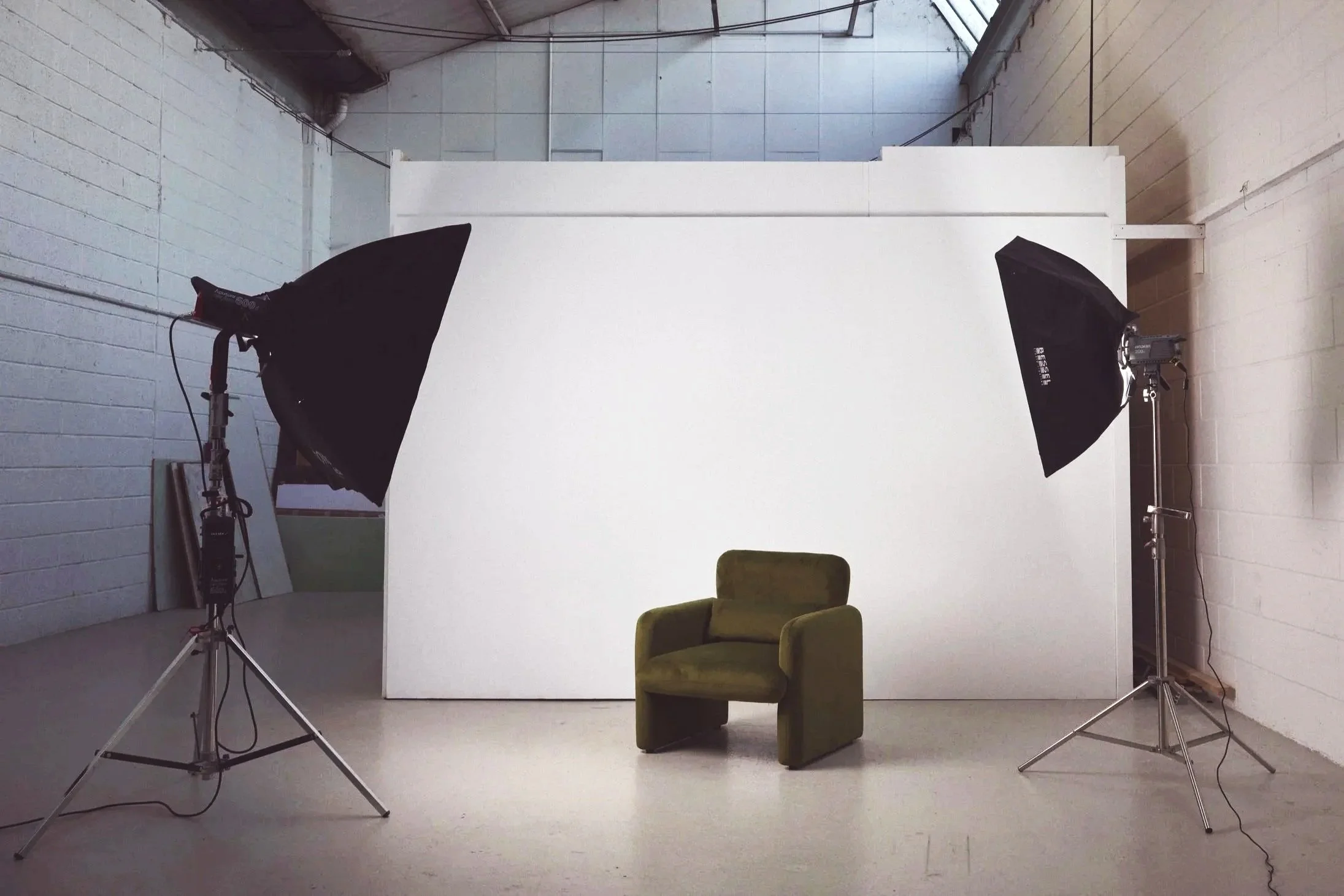 A photography studio setup with two softbox lights directed at a vintage green armchair placed in front of a plain white backdrop, in a large industrial space with exposed ceiling and brick walls.