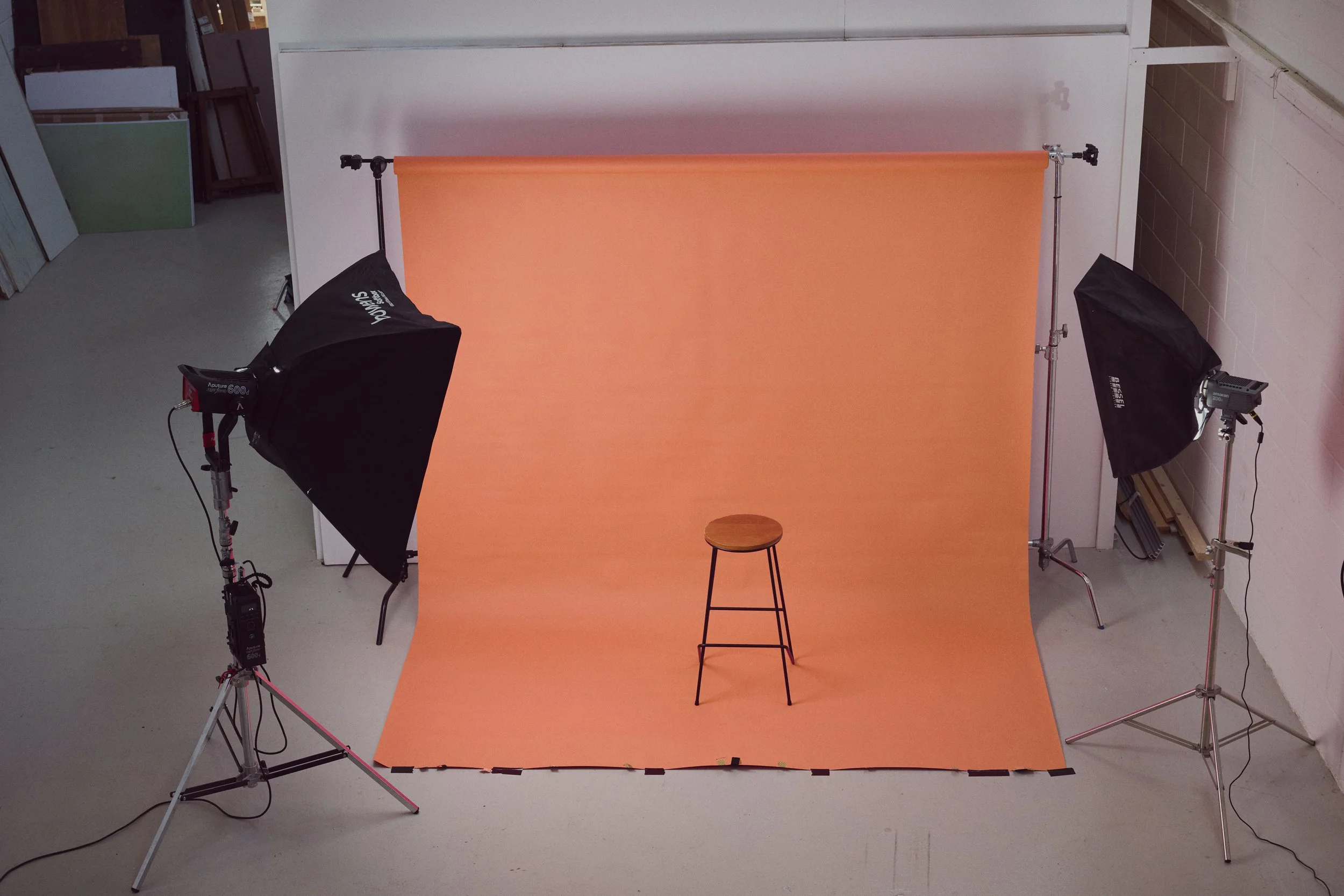 Photography studio setup with a small stool on an orange backdrop, two large softbox lights on tripods, and equipment on the floor in a studio environment.