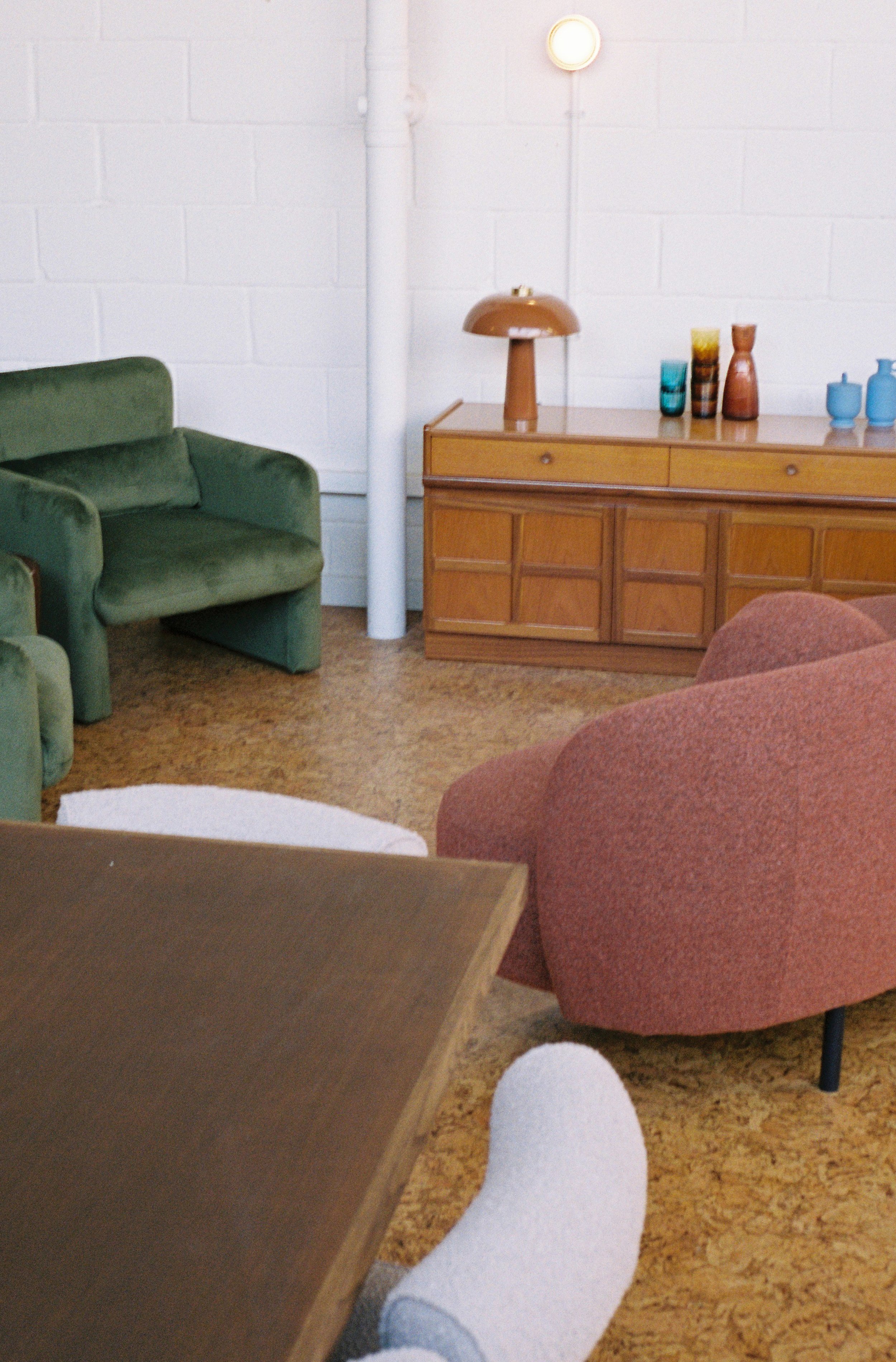 A cozy living room with green and pink armchairs, a wooden sideboard with colorful vases, and a warm lamp, seen from a person's perspective resting their foot on a table.