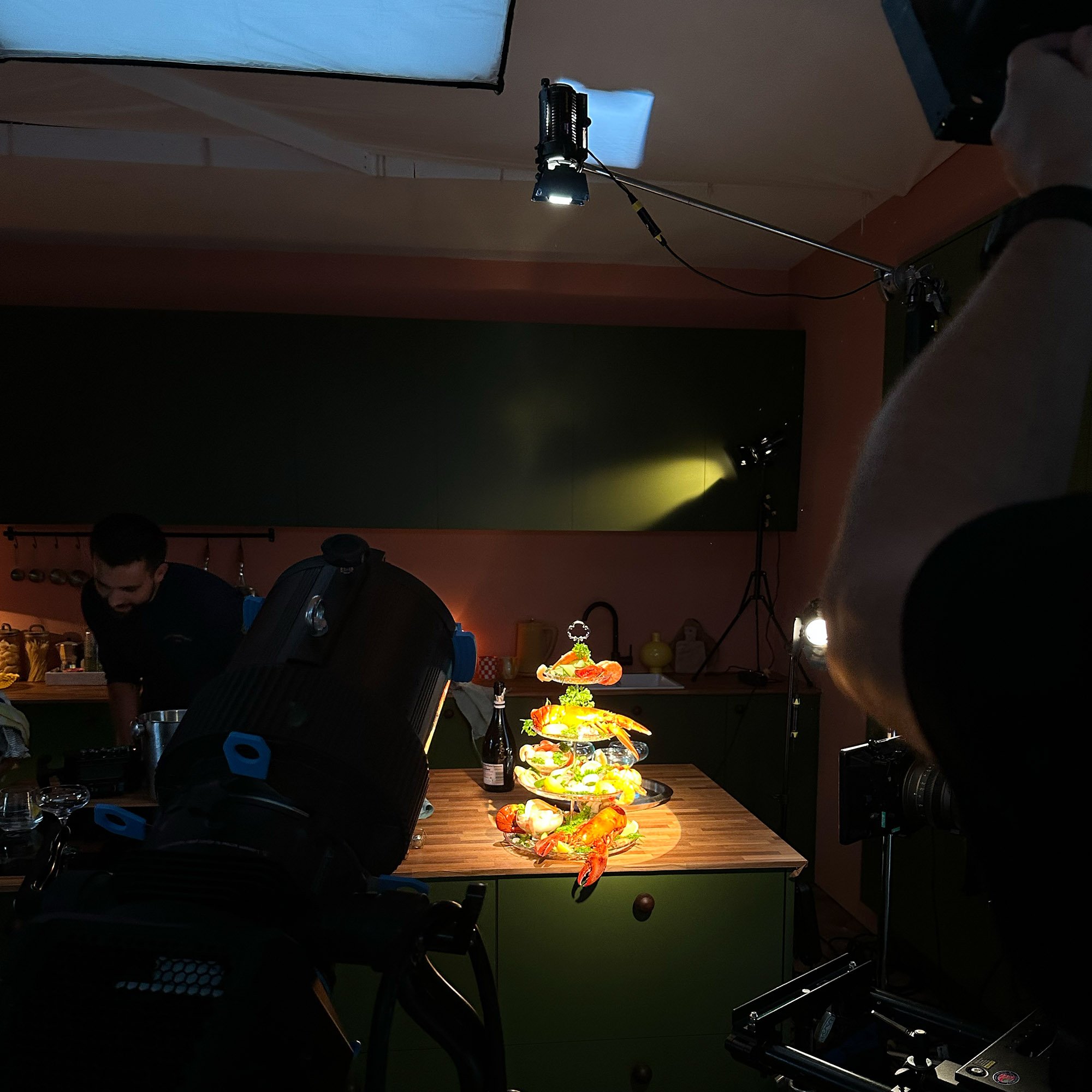 Dark lighting in green and orange kitchen with wooden worktop showing tiered stand of seafood including lobster, king prawns, lettuce. Action shot with camera and lighting
