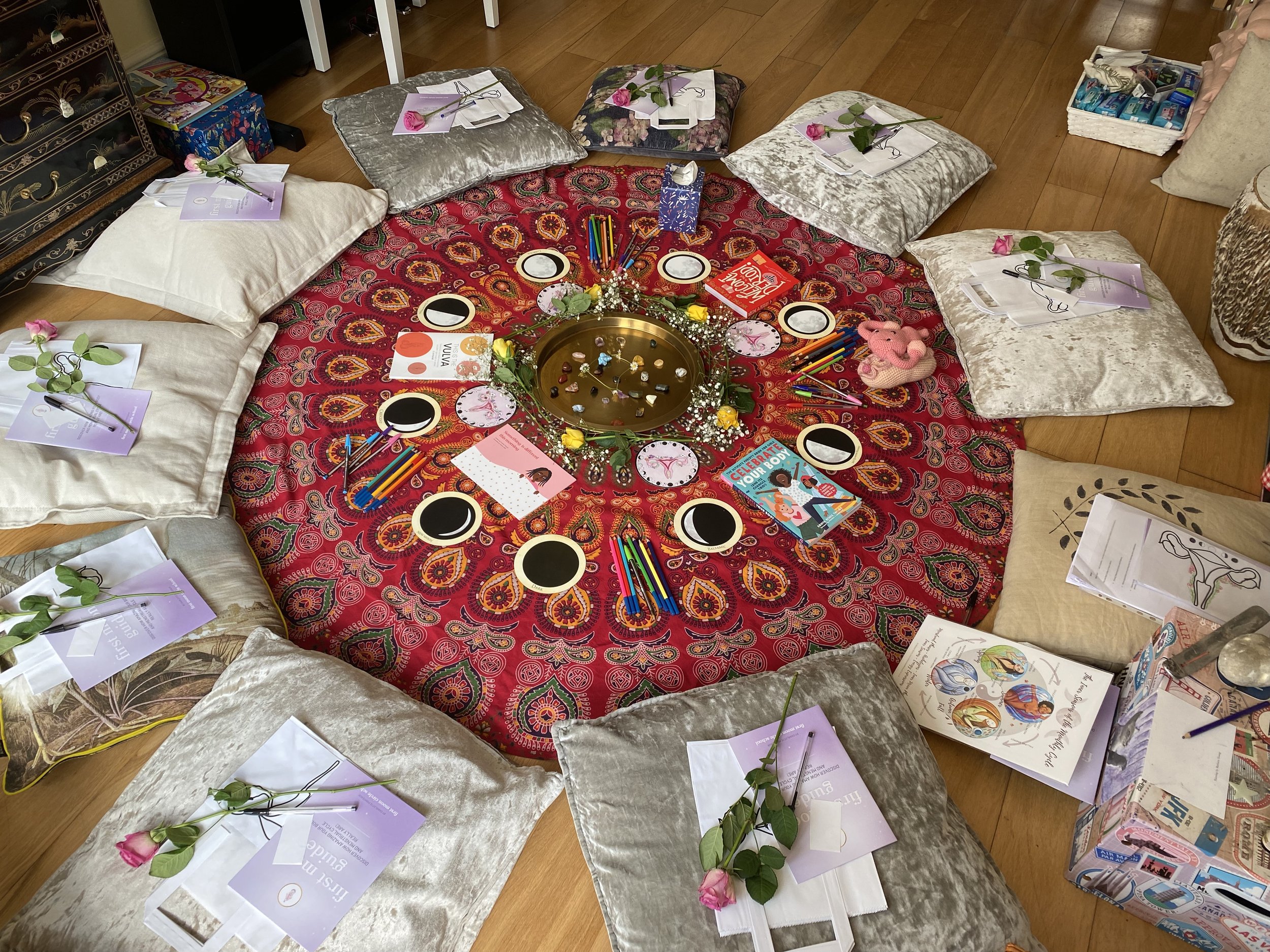 A colorful setup for a gathering with a circular red mandala cloth in the center, surrounded by cushions on a wooden floor. Each cushion has a pink rose and printed materials on top. In the center, there's a gold tray with candles and decorative ston
