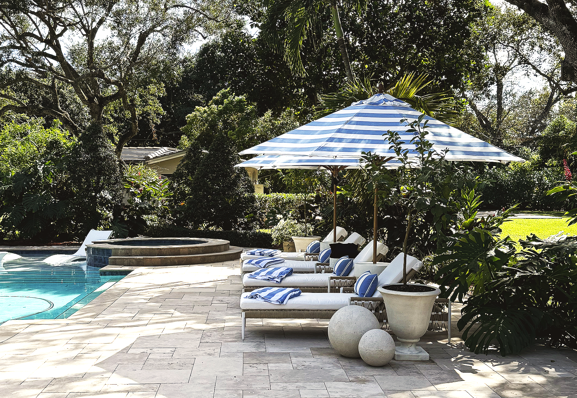 A backyard with a swimming pool, lounge chairs with blue and white striped cushions, a large striped patio umbrella, and lush greenery with tall trees and bushes surrounding the area.