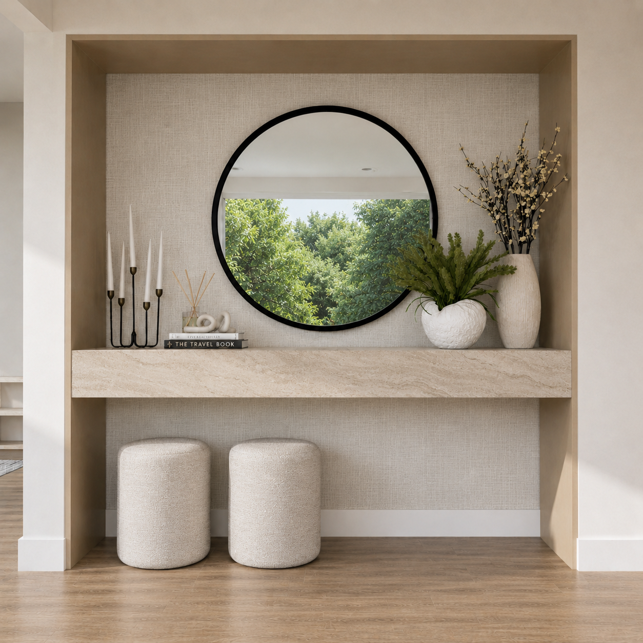 Decorative built-in wall niche with a round mirror, two vases with greenery and branches, a candle holder with five white candles, two books, and decorative objects, with two beige ottomans in front on a wooden floor.