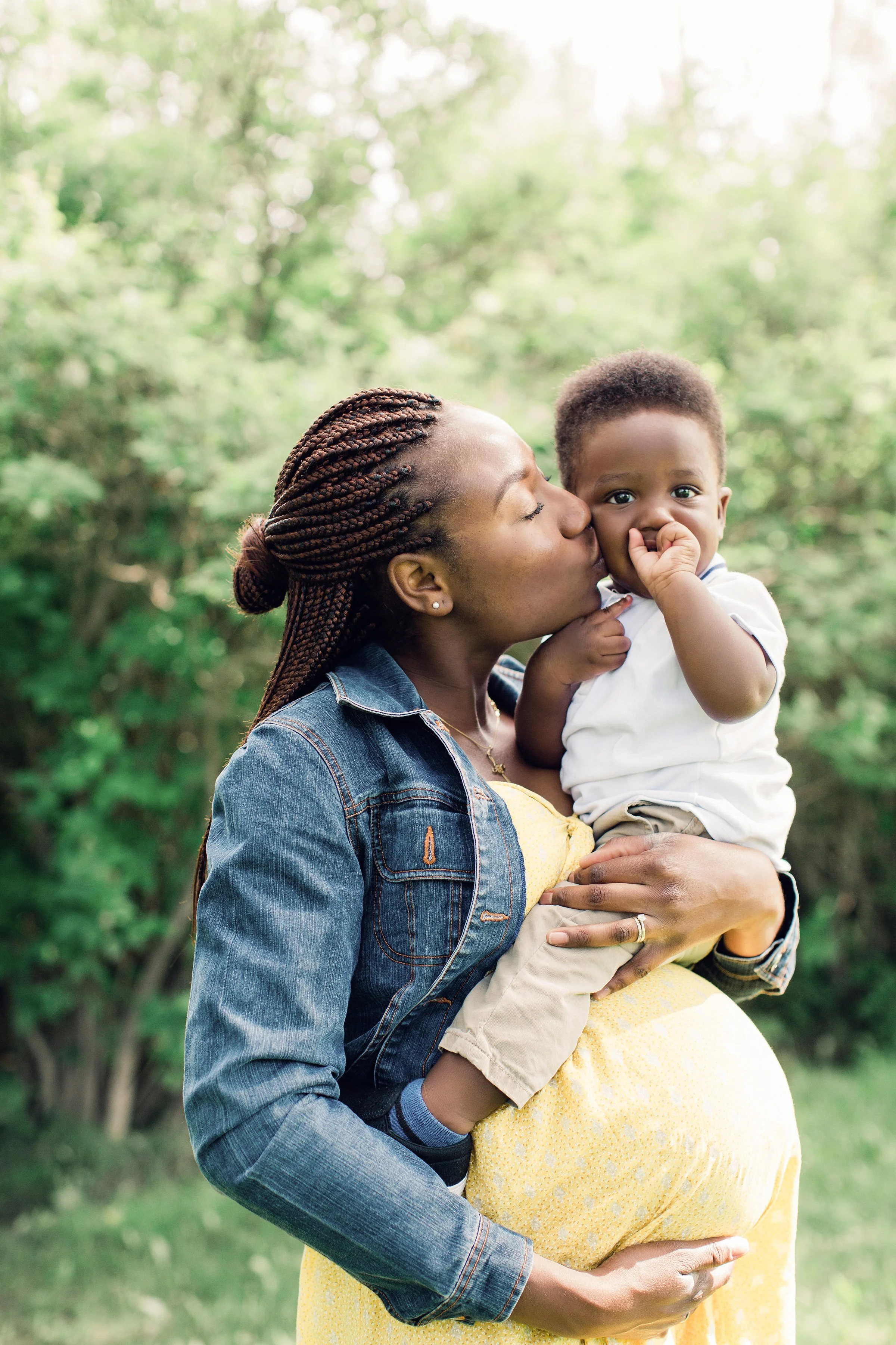 A woman with braided hair wearing a denim jacket and yellow dress holding a young child, giving him a kiss on the cheek outdoors with green trees in the background.
