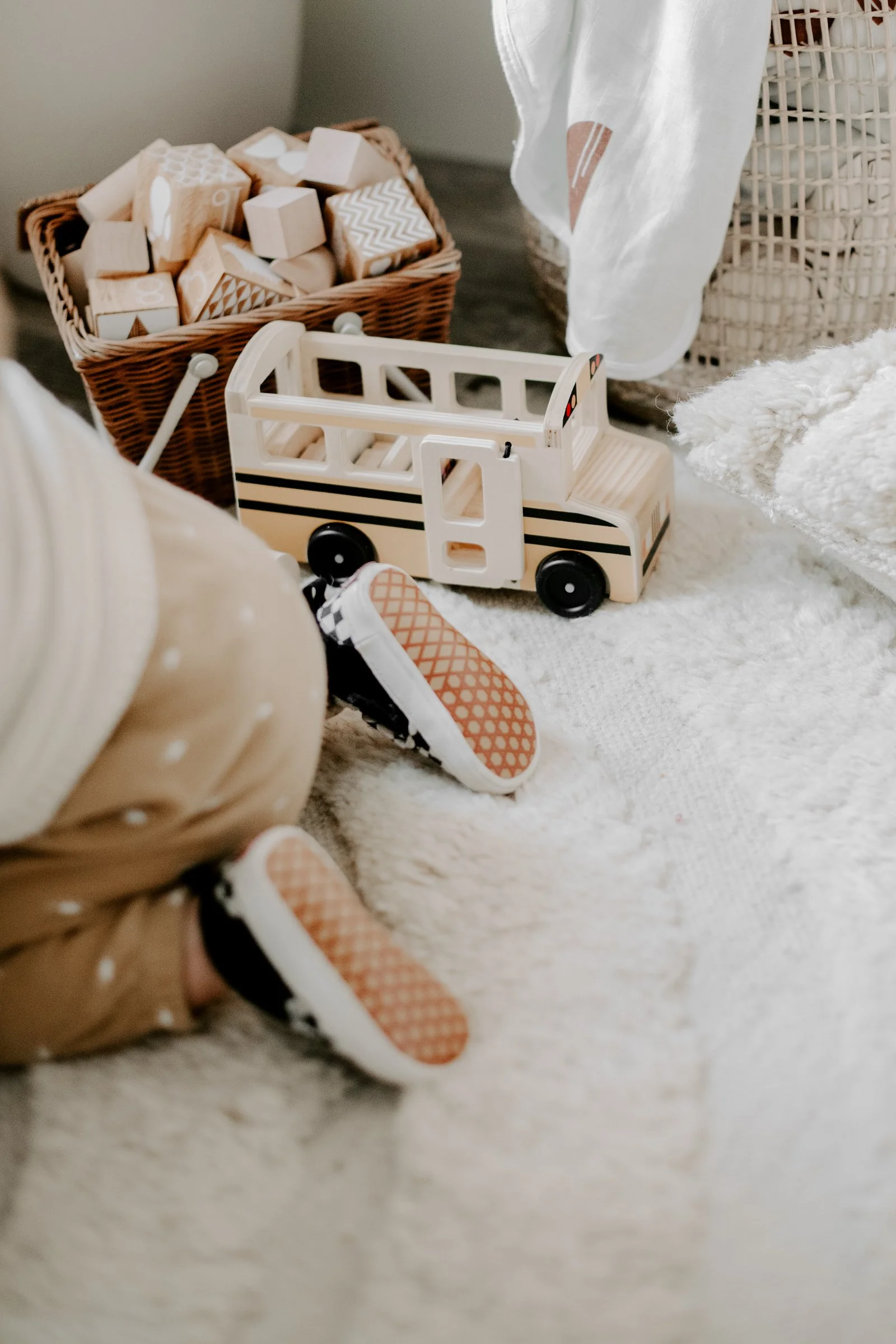 Children's toy scene with a white wooden bus, a basket of wooden blocks, a sneaker, a carpeted floor, and some fabric and wicker baskets.