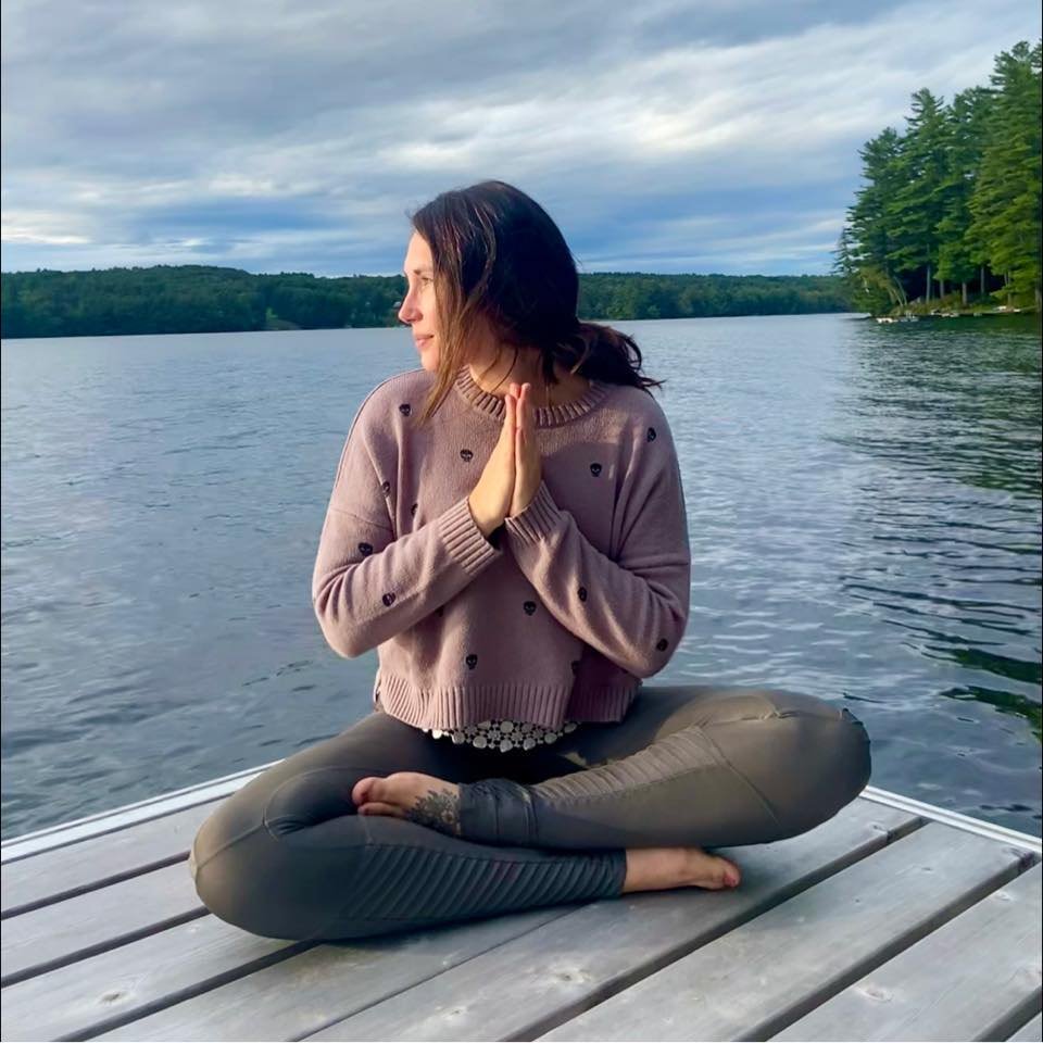 A woman sitting cross-legged on a wooden dock by a lake, with her hands in a prayer position and eyes closed, during cloudy weather surrounded by trees.