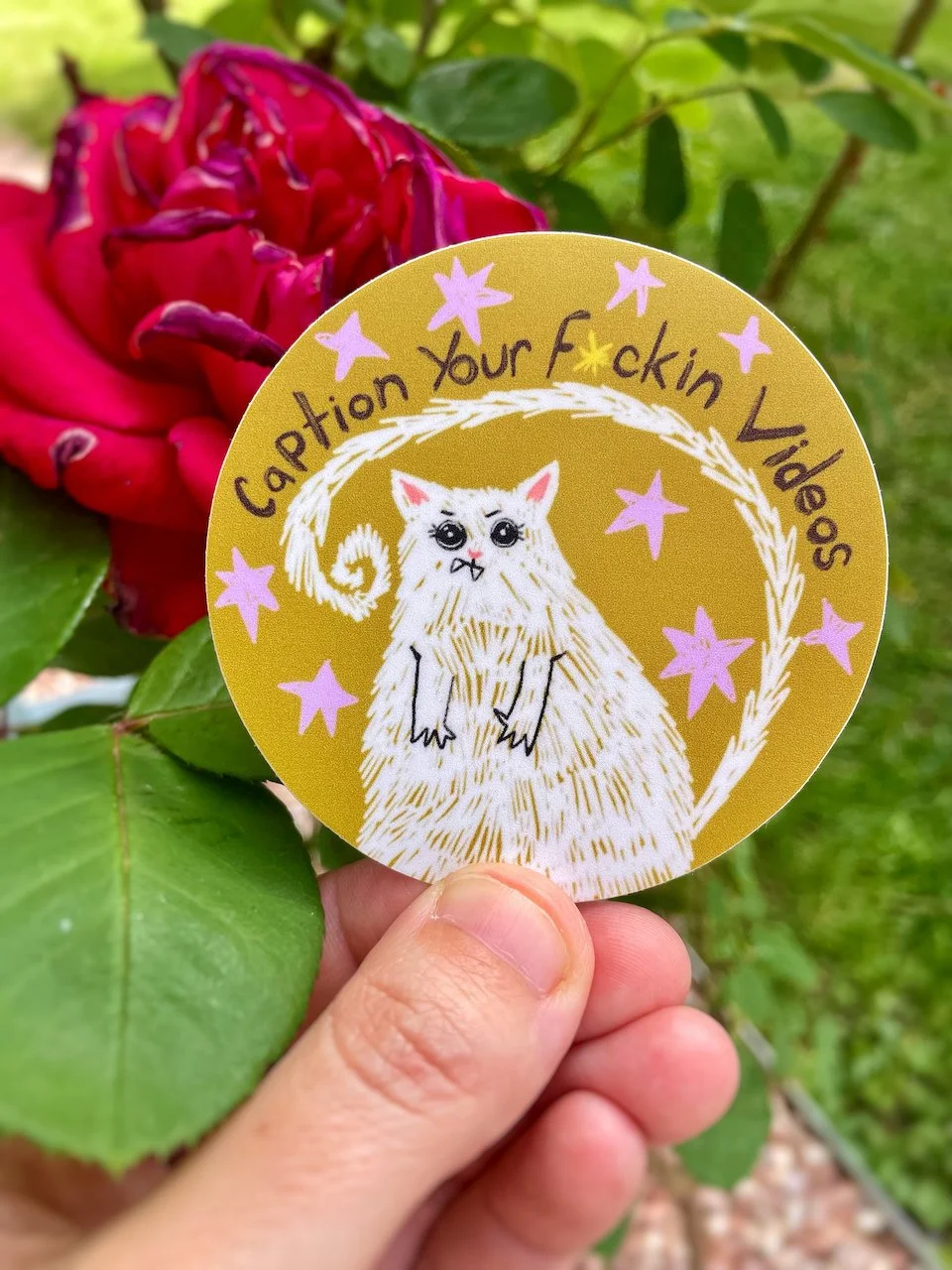 a round sticker of a illustrated white cat with words above it reading 'caption your f*ckin videos'