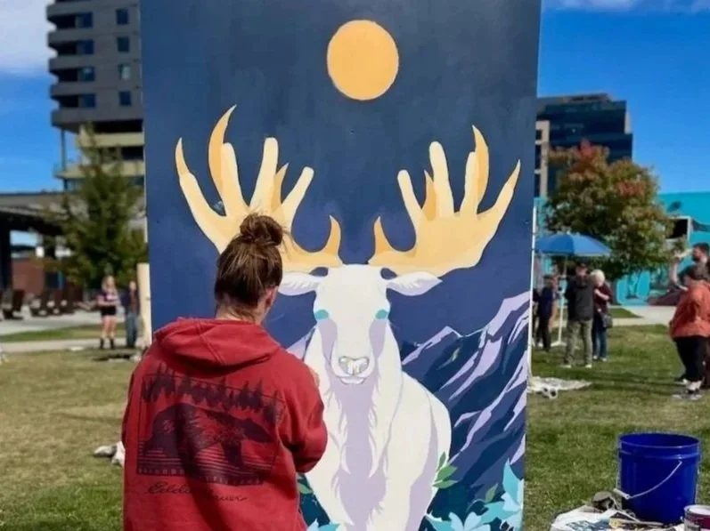 An artist stands in front of her mural panel which shows an elk under a moon.