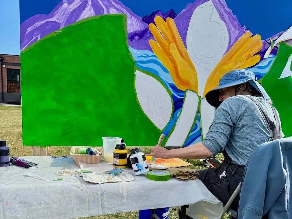 An artist sits in front of her mural panel in a chair with a table. She is adding paint to a paintbrush.