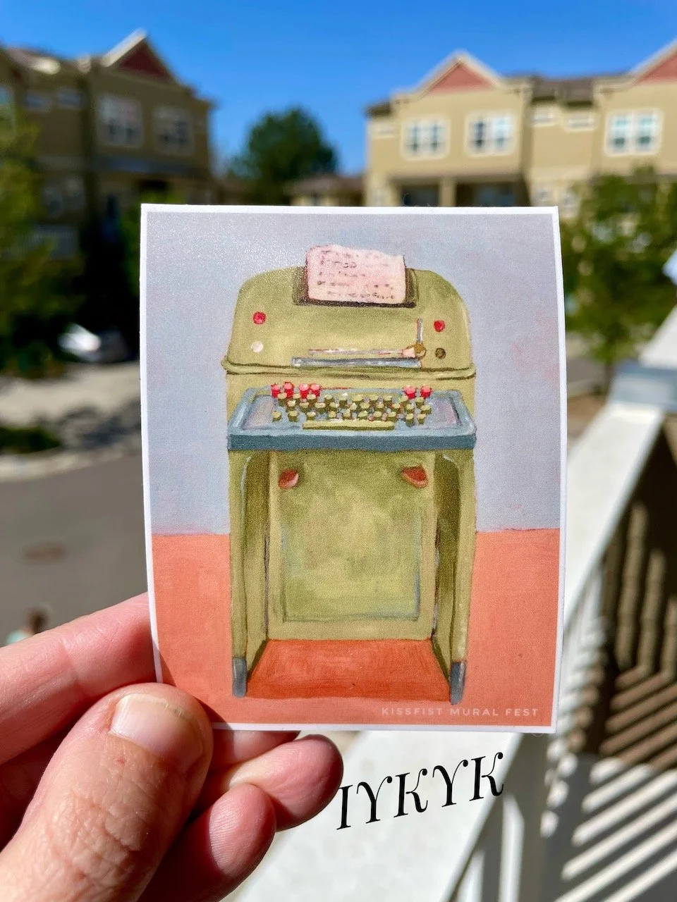 a sticker of a green monster old fashioned TTY held in a white hand in an outdoor neighborhood environment. Text on screen reads IYKYK.