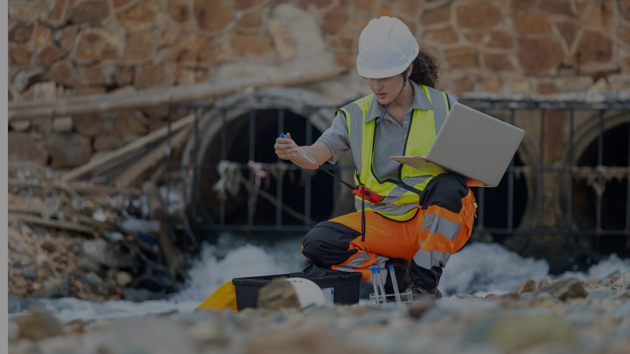A female scientist in safety gear taking water samples at a river site.