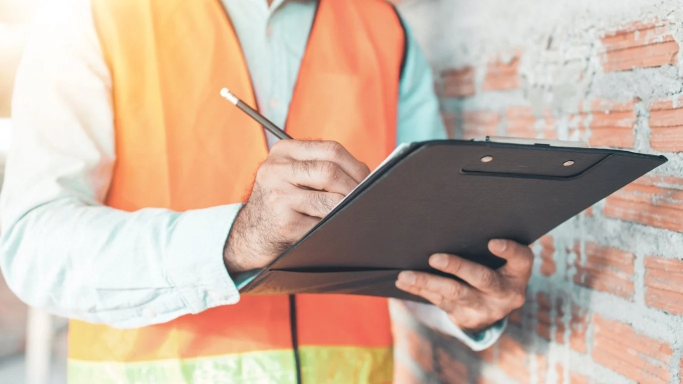 A person wearing an orange safety vest and light blue shirt, holding a tablet and writing on it with a pencil, standing against a brick wall.