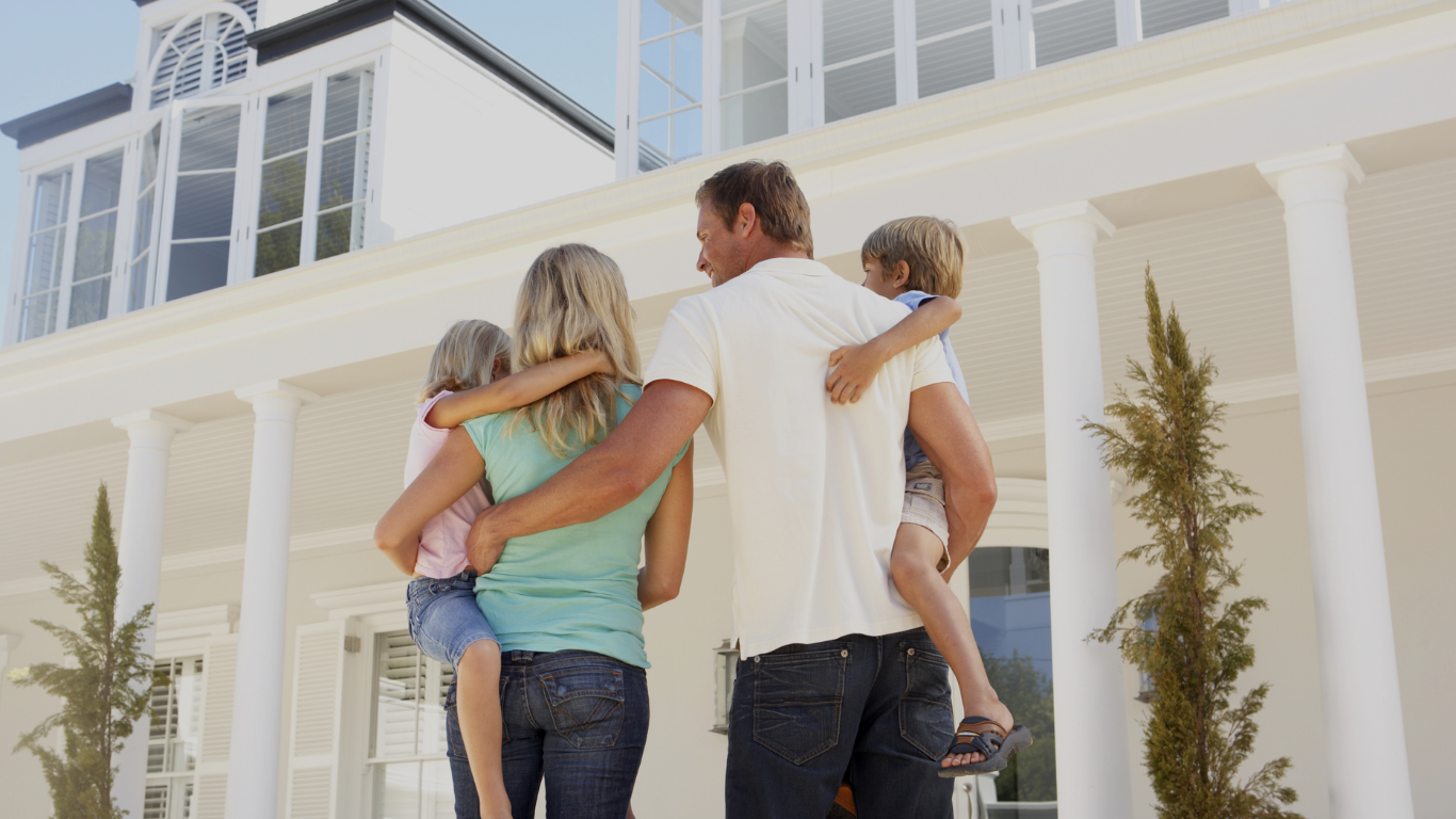 A family of four, two adults and two children, standing in front of a large white house with tall pillars, embracing and smiling.