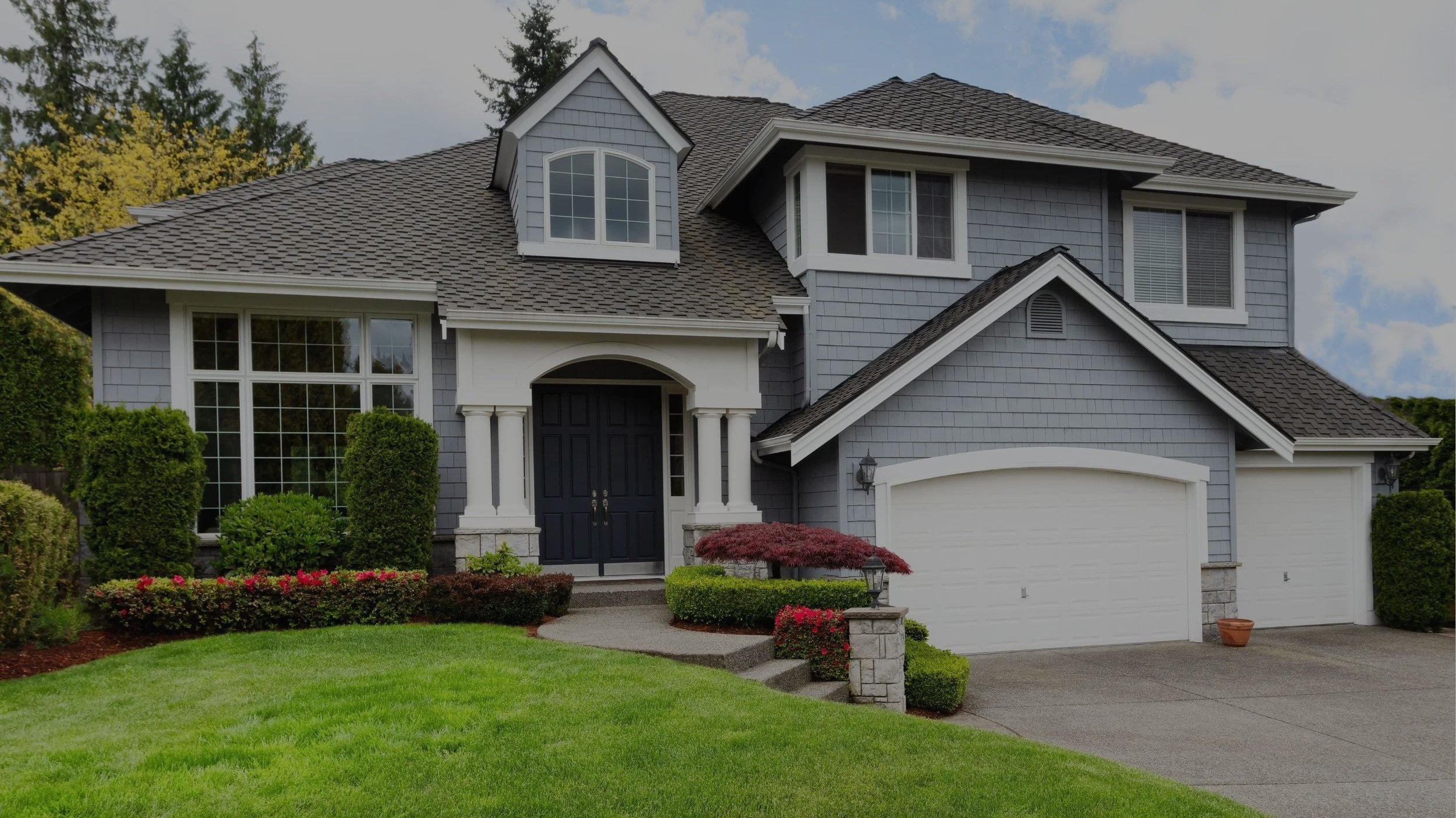 A two-story gray house with white trim and a dark front door, surrounded by a neatly landscaped lawn and garden with bushes, small trees, and flowers, under a partly cloudy sky.
