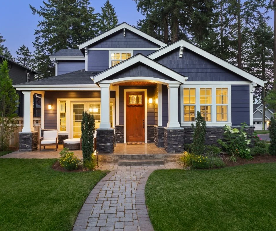 Front view of a modern two-story house with a blue exterior, white trim, and large windows. The house has a covered porch with white columns, outdoor seating, and a wooden front door. The yard features a stone walkway, green grass, and landscaped plants, with tall trees in the background.